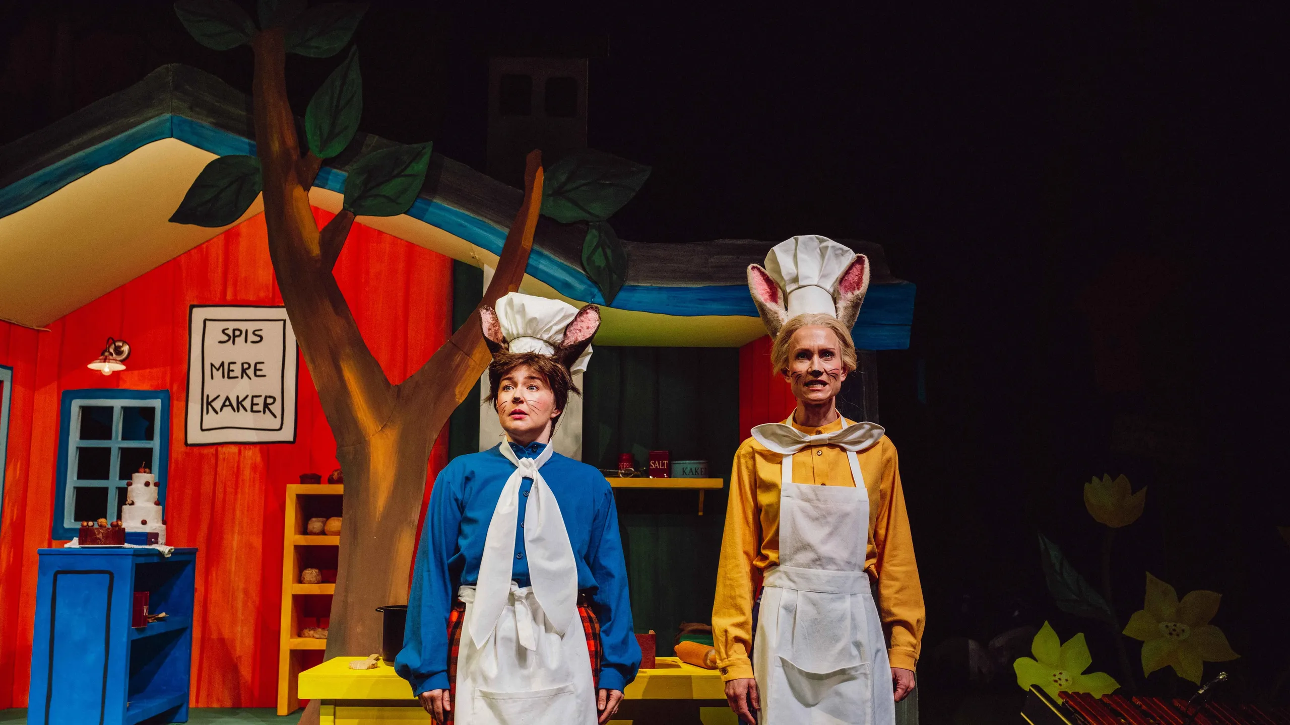 To bakere med museører står foran et fargerikt hus med skiltet 'Spis mere kaker'.