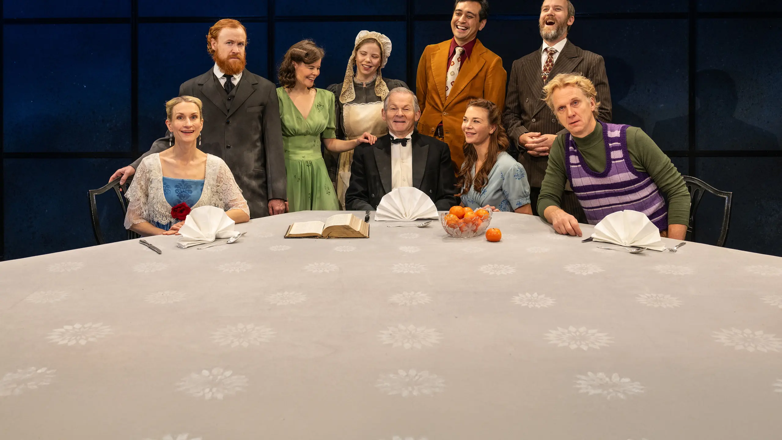 Ni skuespillere i kostymer fra forskjellige tidsaldre ved et pyntet bord