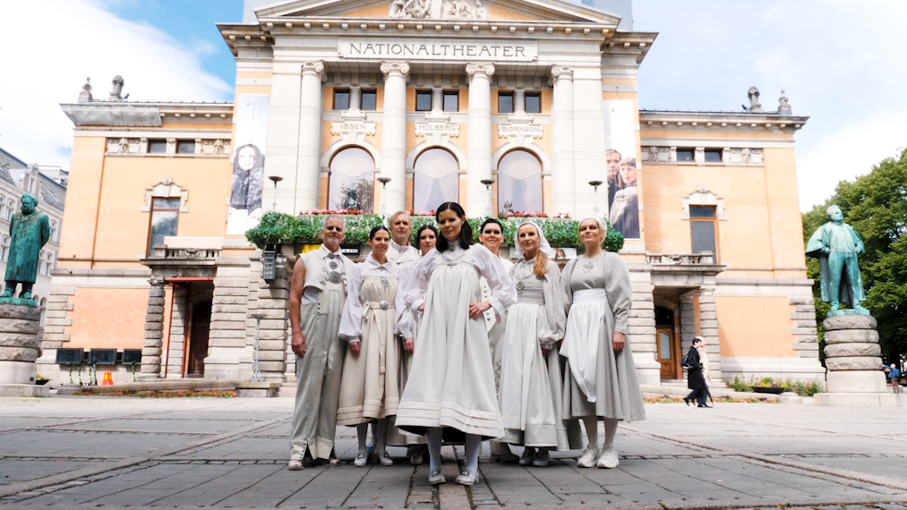 Skuespillere i hvite bunadaktige kostymer foran Nationaltheatret