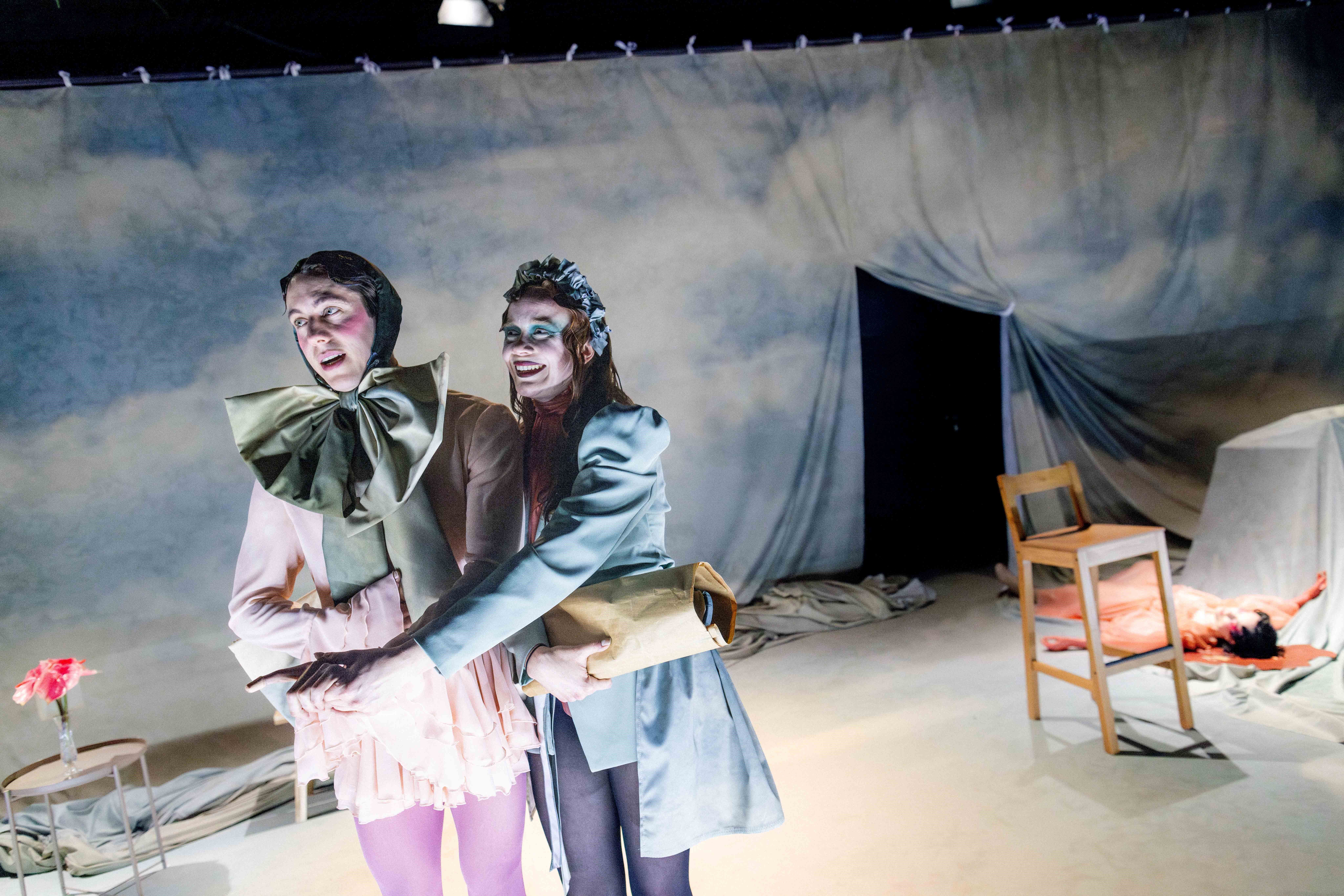 To personer i kostymer står sammen i et scenelandskap; en peker fremover mens den andre holder en pakke