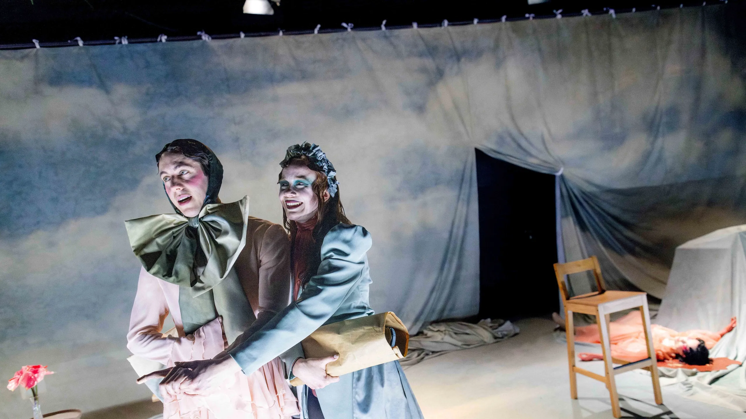 To personer i kostymer står sammen i et scenelandskap; en peker fremover mens den andre holder en pakke