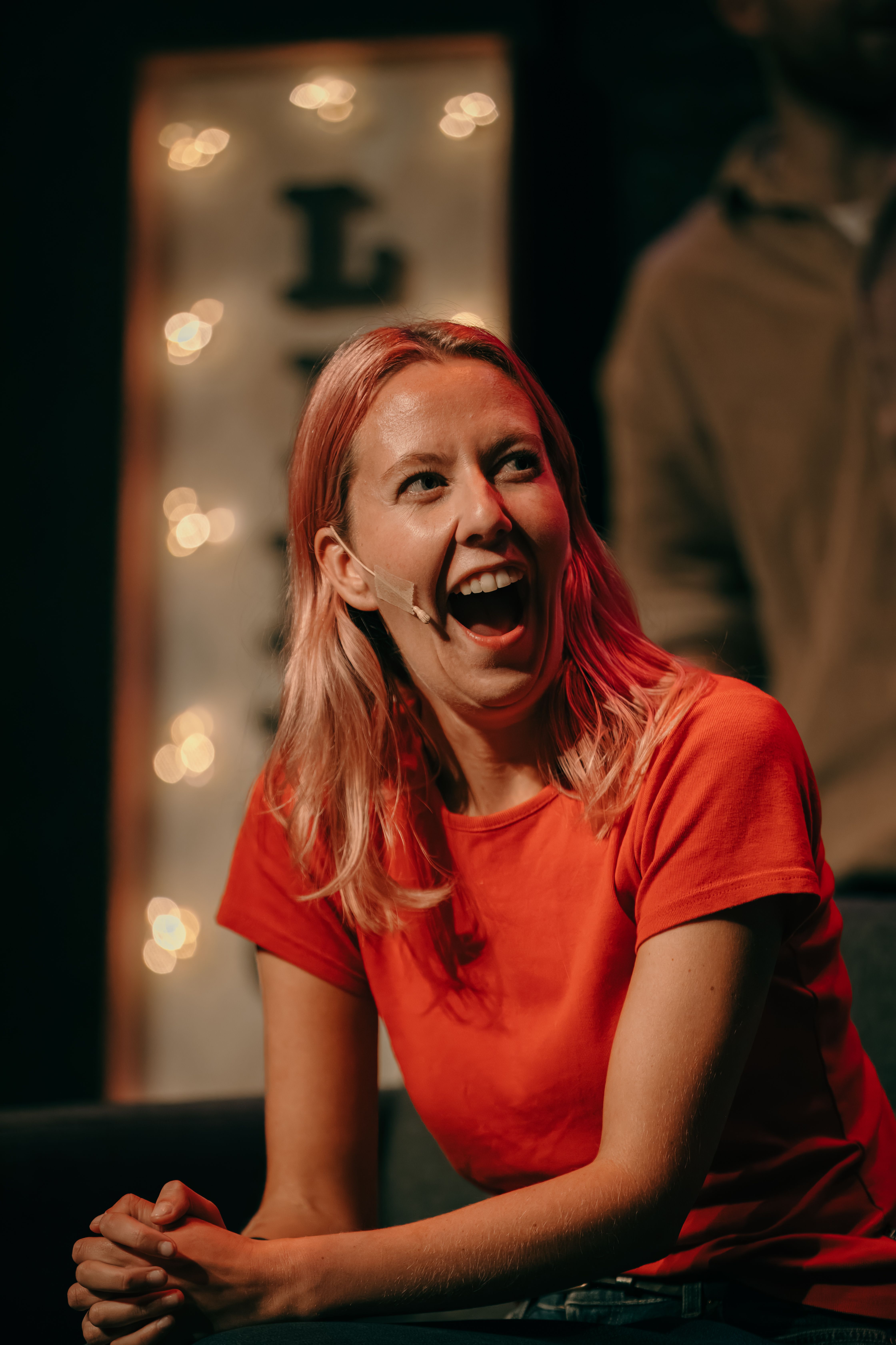 Nærbilde av person i rød t-skjorte med mikrofon på kinnet som ler med åpen munn