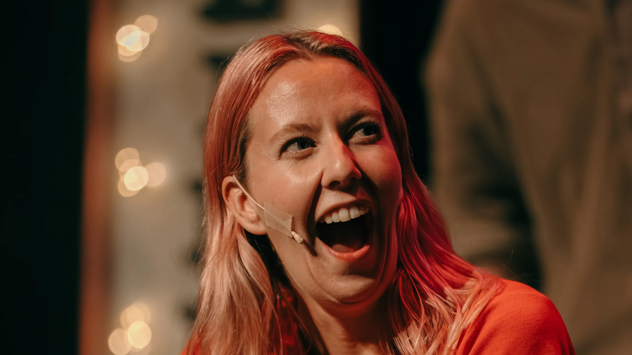 Nærbilde av person i rød t-skjorte med mikrofon på kinnet som ler med åpen munn