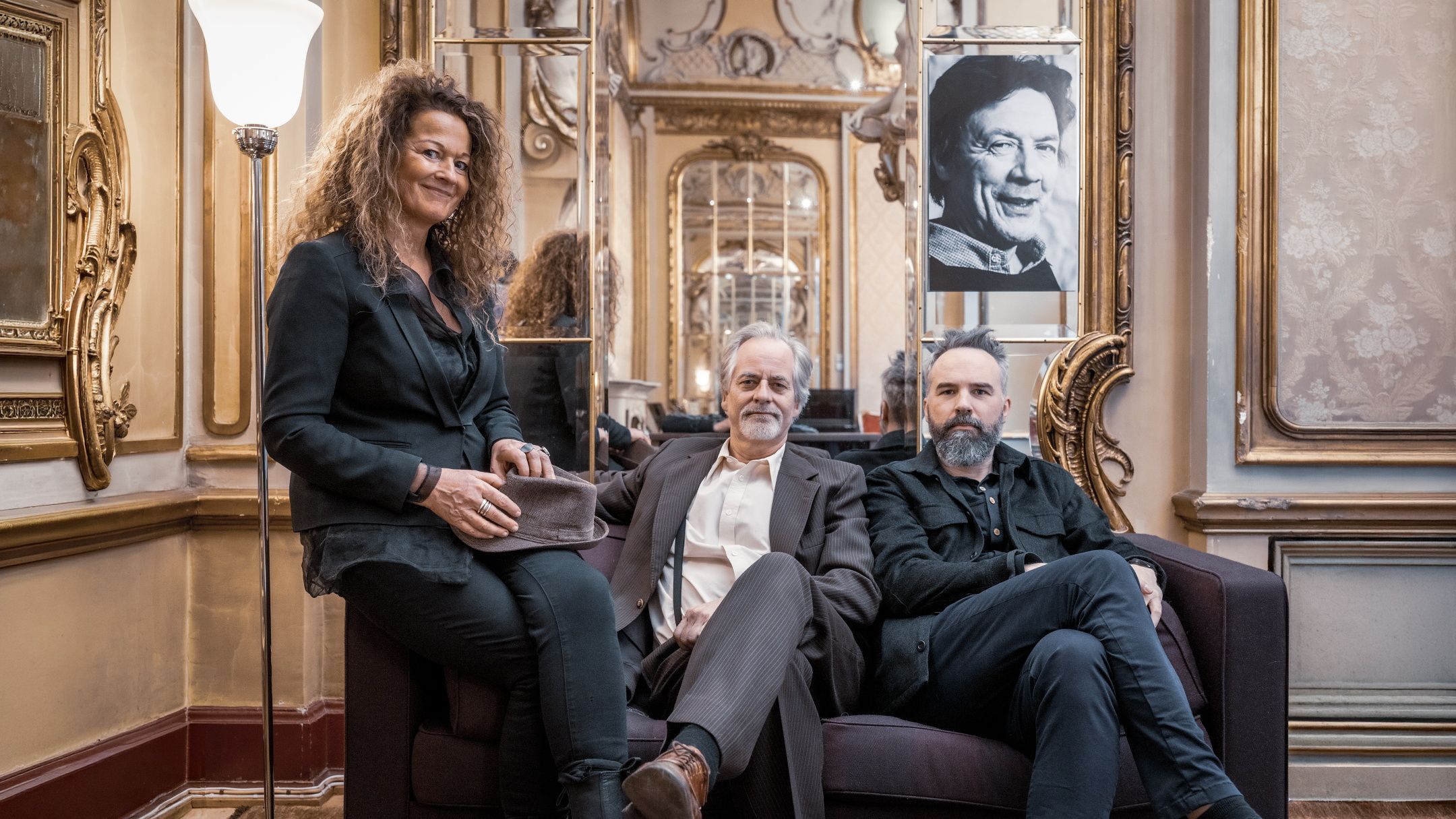 Tre mennesker sitter i en sofa i et gullforgylt rom og ser mot kameraet