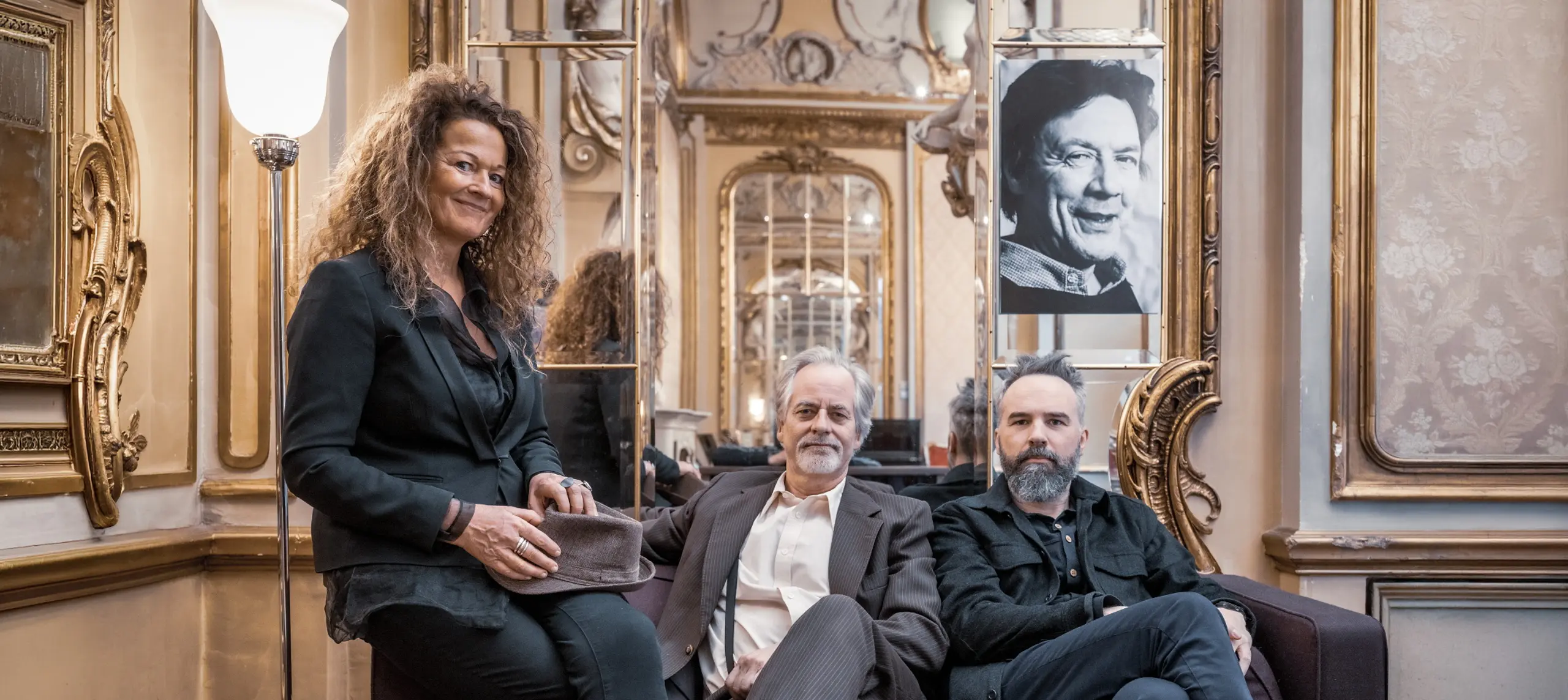 Tre mennesker sitter i en sofa i et gullforgylt rom og ser mot kameraet