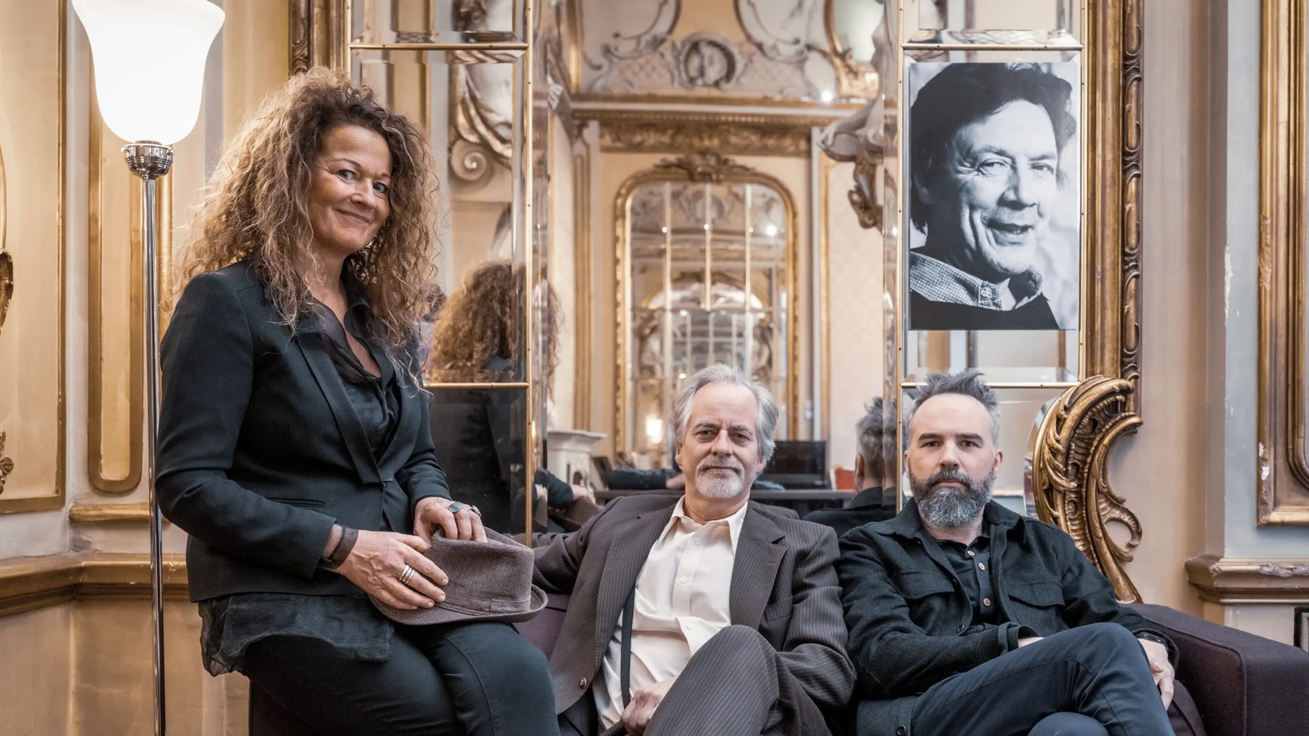 Tre mennesker sitter i en sofa i et gullforgylt rom og ser mot kameraet