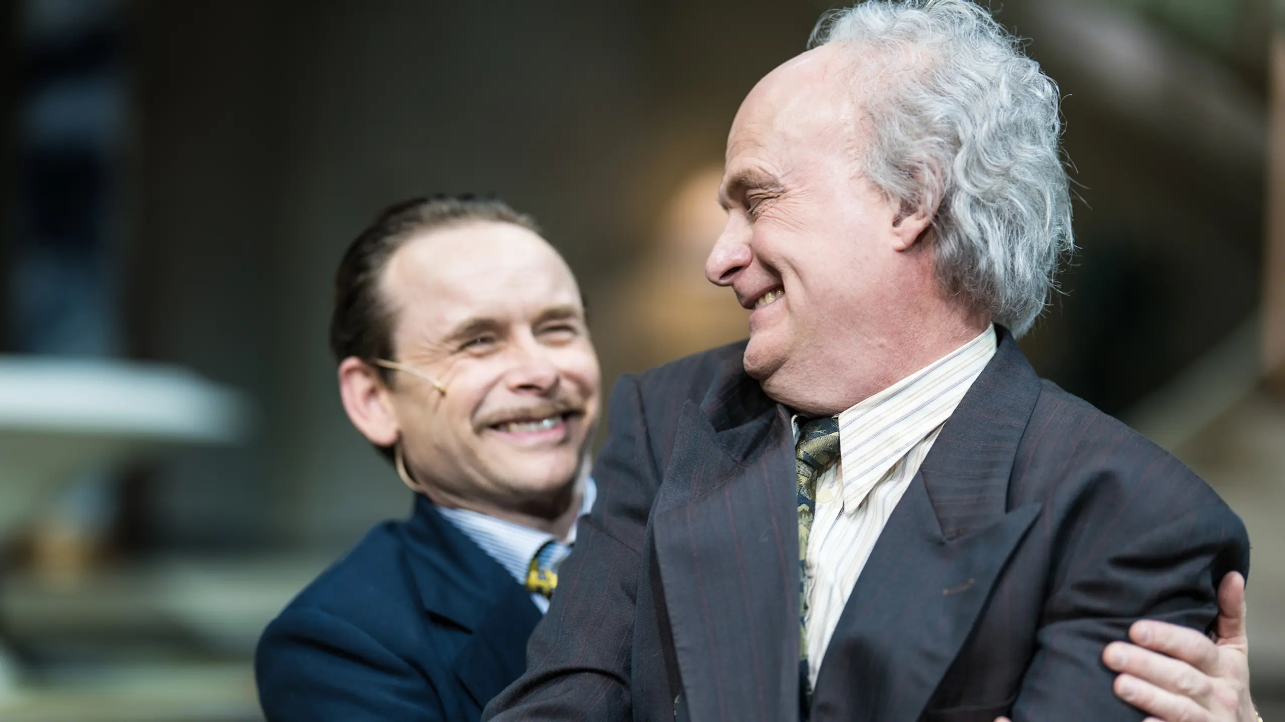 To menn i dress smiler og omfavner hverandre på teaterscene