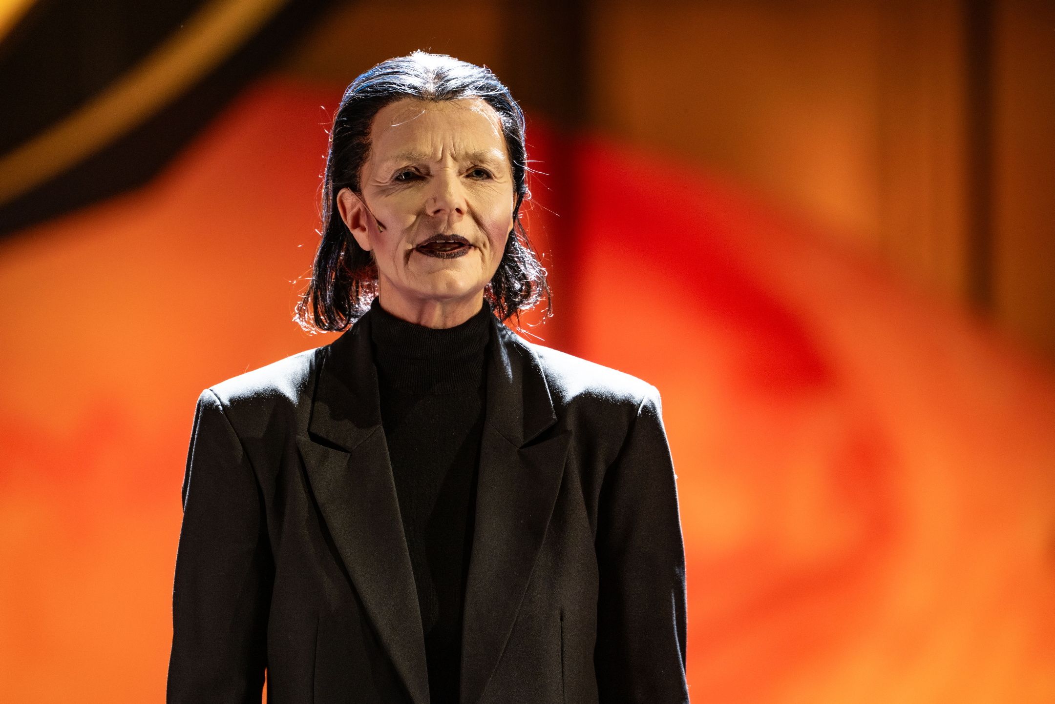 Person med sort hår og teatersminke iført svart dress står alvorlig foran rød-oransje bakgrunn.