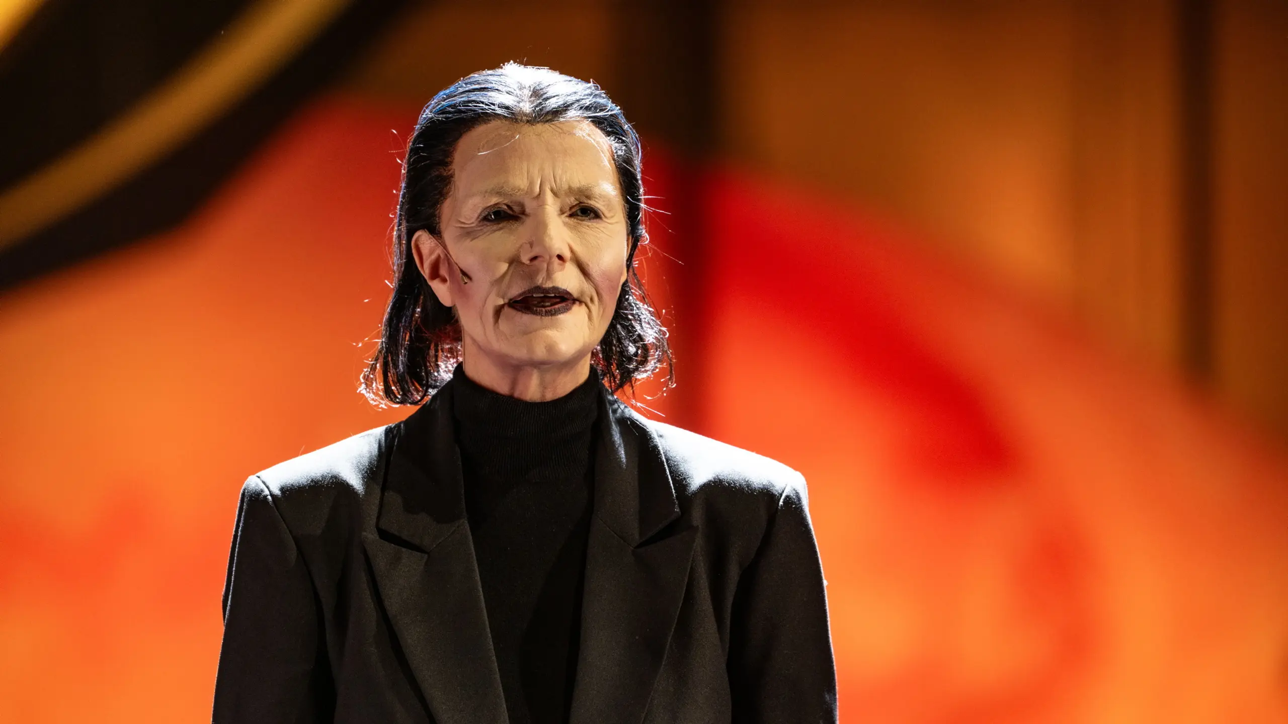 Person med sort hår og teatersminke iført svart dress står alvorlig foran rød-oransje bakgrunn.