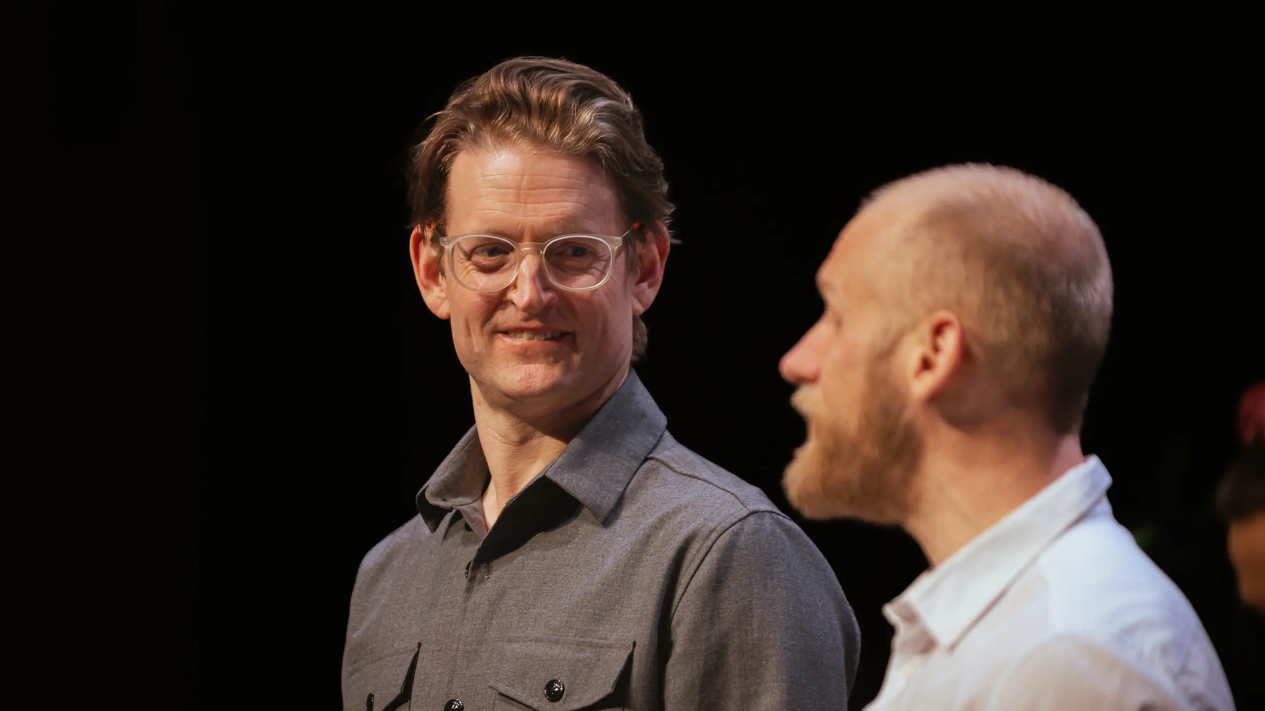 To personer i samtale, én med briller smiler mens den andre snakker i profil