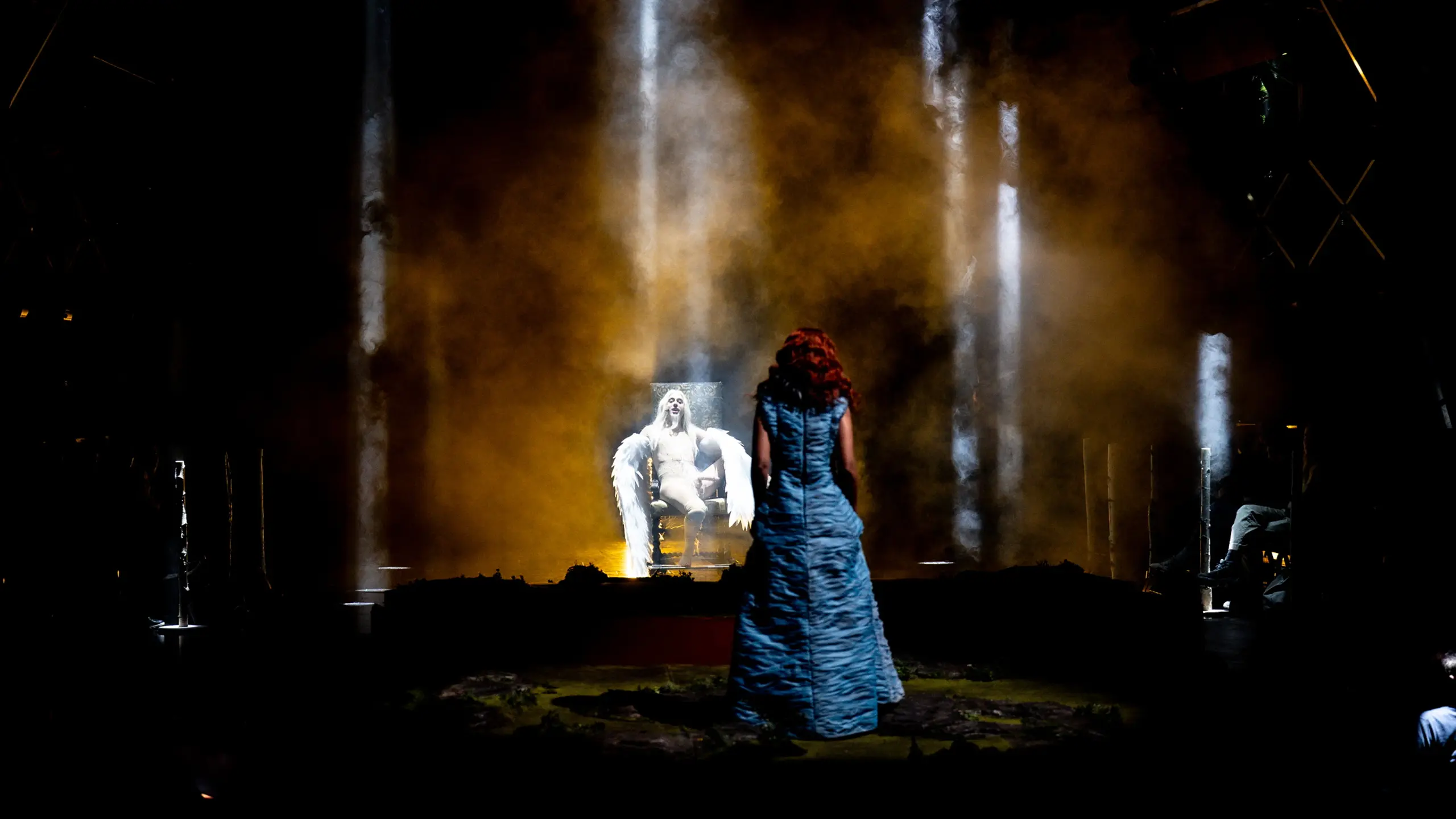 En figur med hvite vinger og kroppsmaling sitter på en trone omgitt av røyk mens en person i blå kjole står foran.