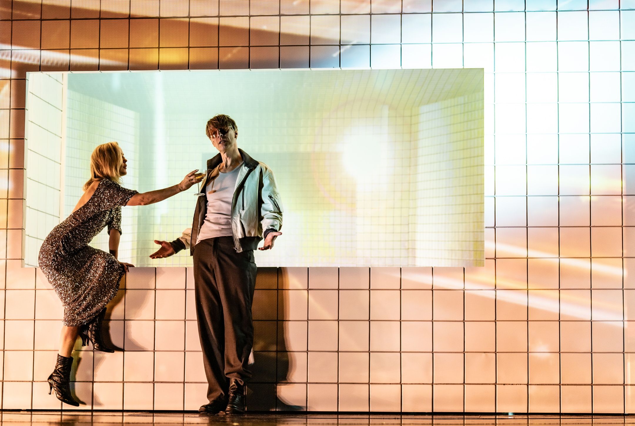 Kvinne i paljettkjole lener seg mot mann i hvit jakke foran lysprojeksjon på flislagt vegg