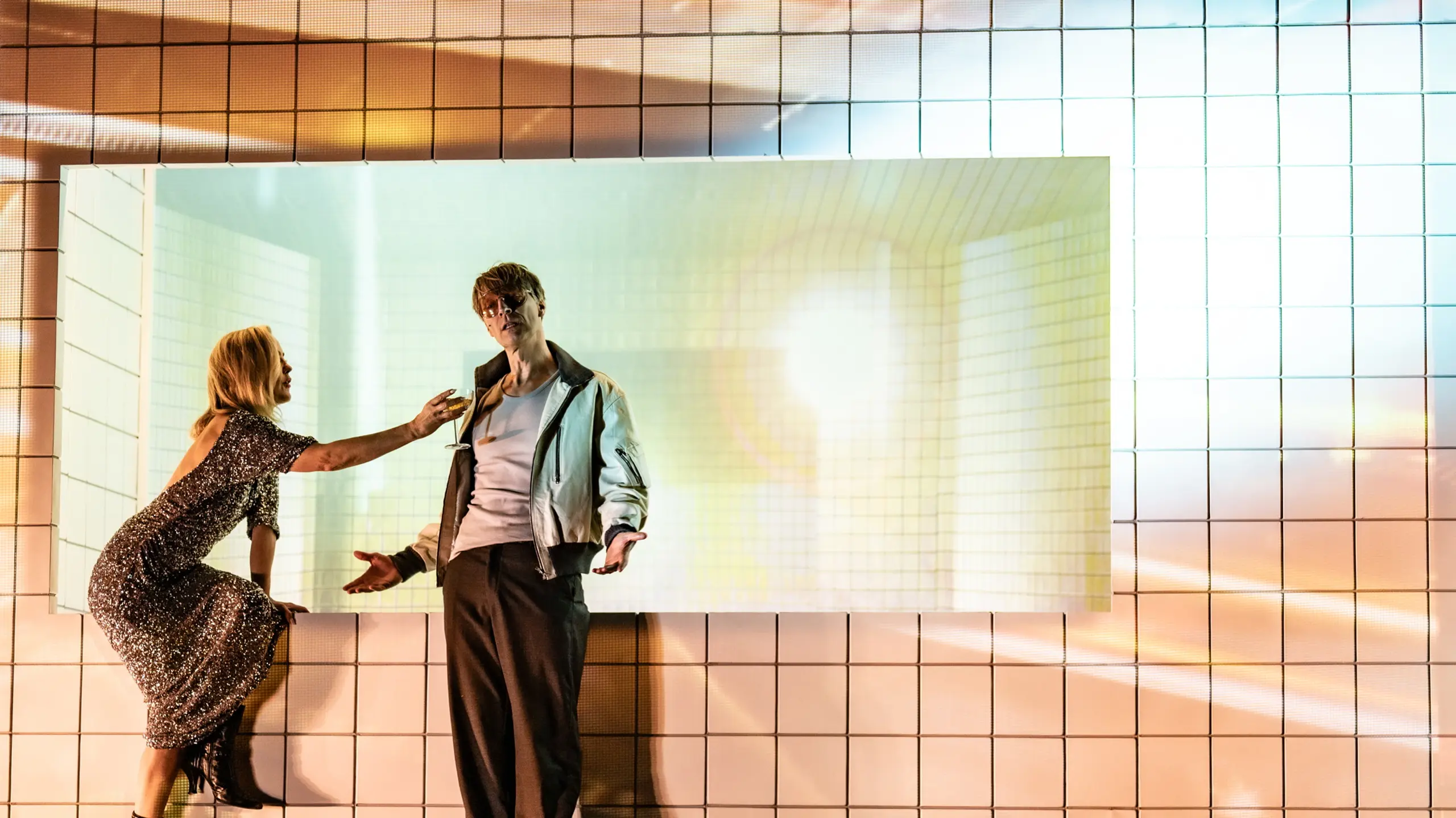 Kvinne i paljettkjole lener seg mot mann i hvit jakke foran lysprojeksjon på flislagt vegg