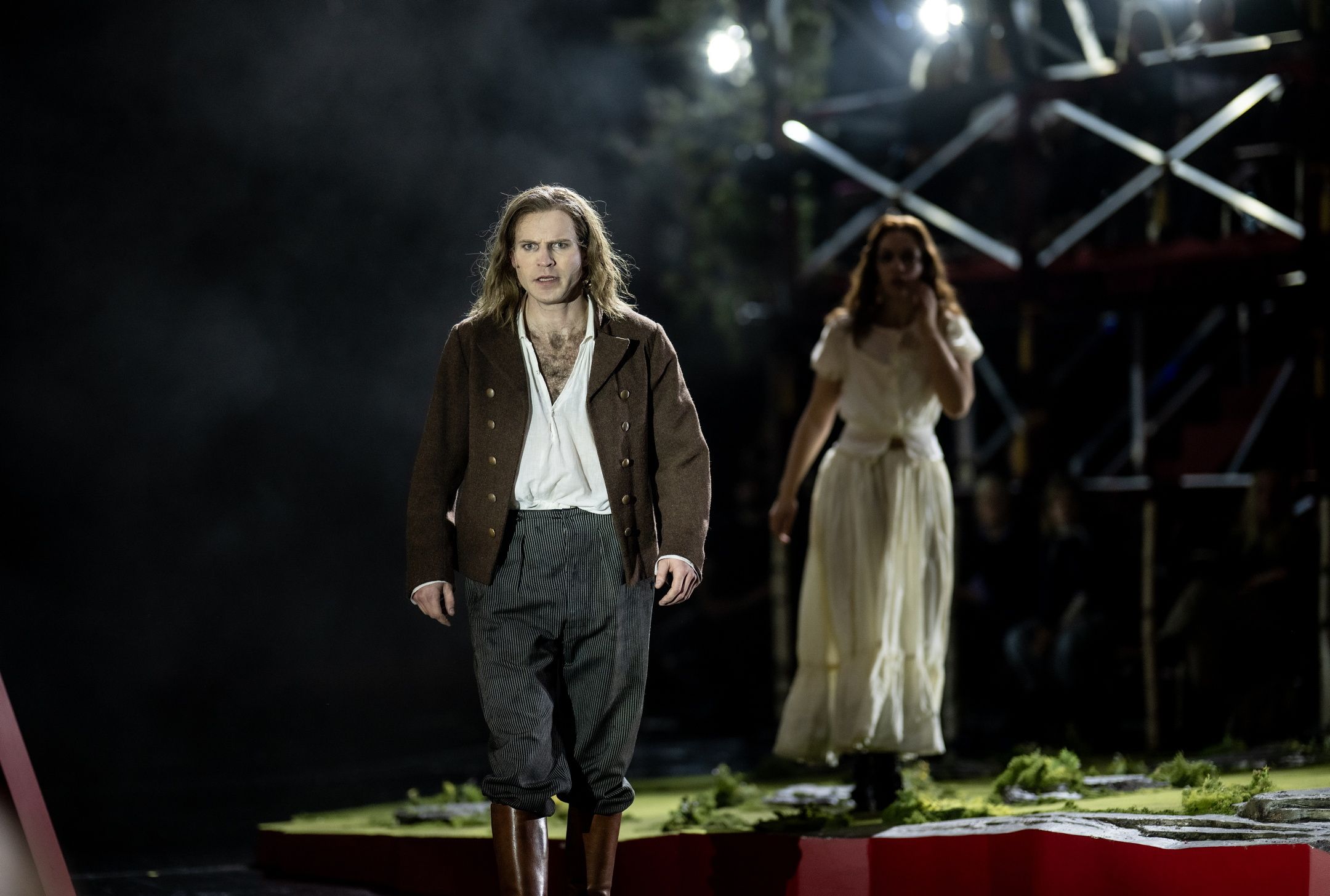 En mann i brune klær og støvler står fremst på scenen med et intenst blikk, mens en kvinne følger ham i bakgrunnen.