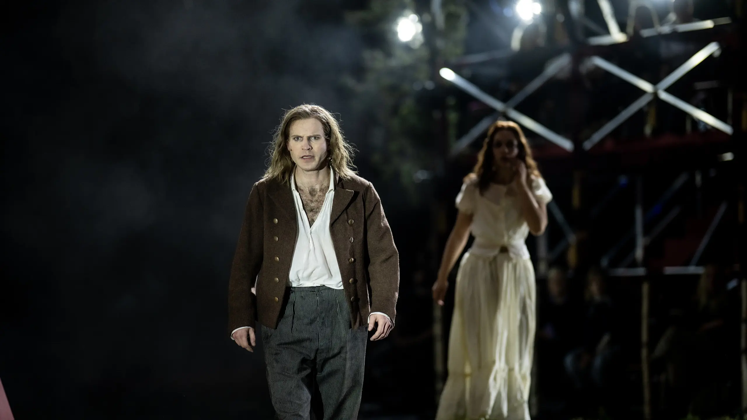 En mann i brune klær og støvler står fremst på scenen med et intenst blikk, mens en kvinne følger ham i bakgrunnen.