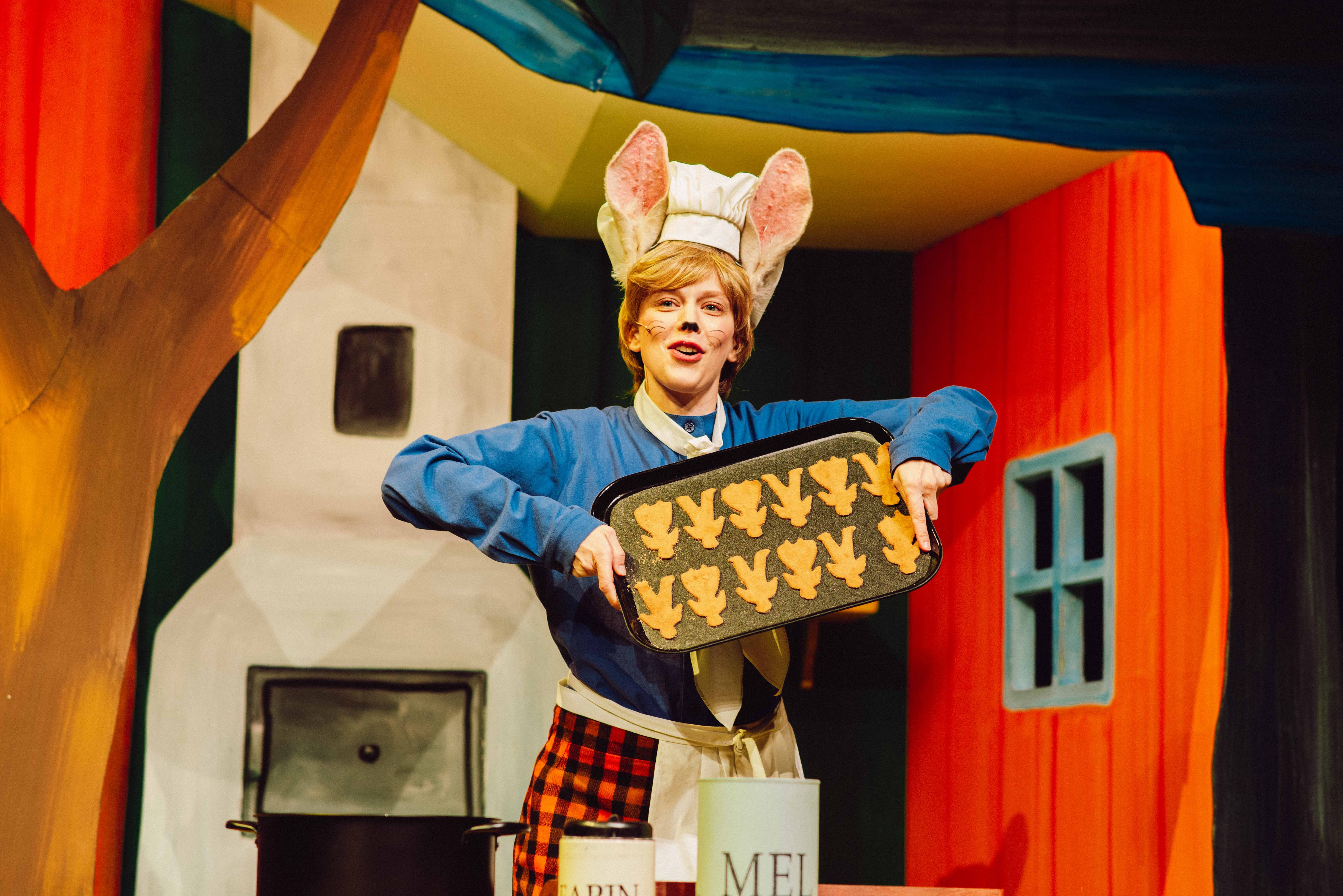 En skuespiller utkledd som hare med store ører og kokkelue holder et brett med småkaker formet som mus, i et fargerikt scenemiljø.