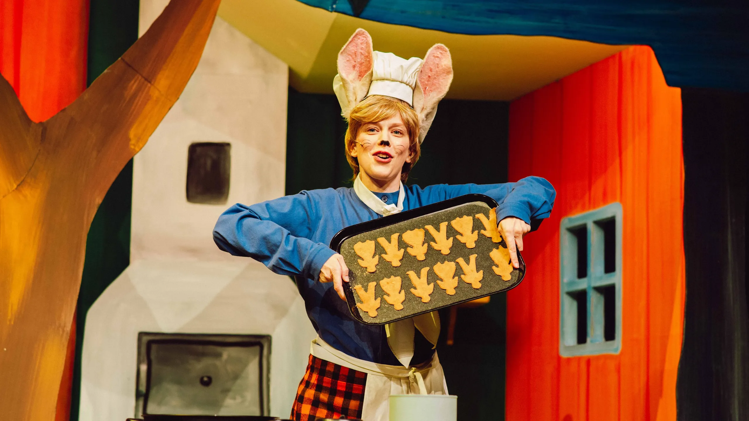 En skuespiller utkledd som hare med store ører og kokkelue holder et brett med småkaker formet som mus, i et fargerikt scenemiljø.