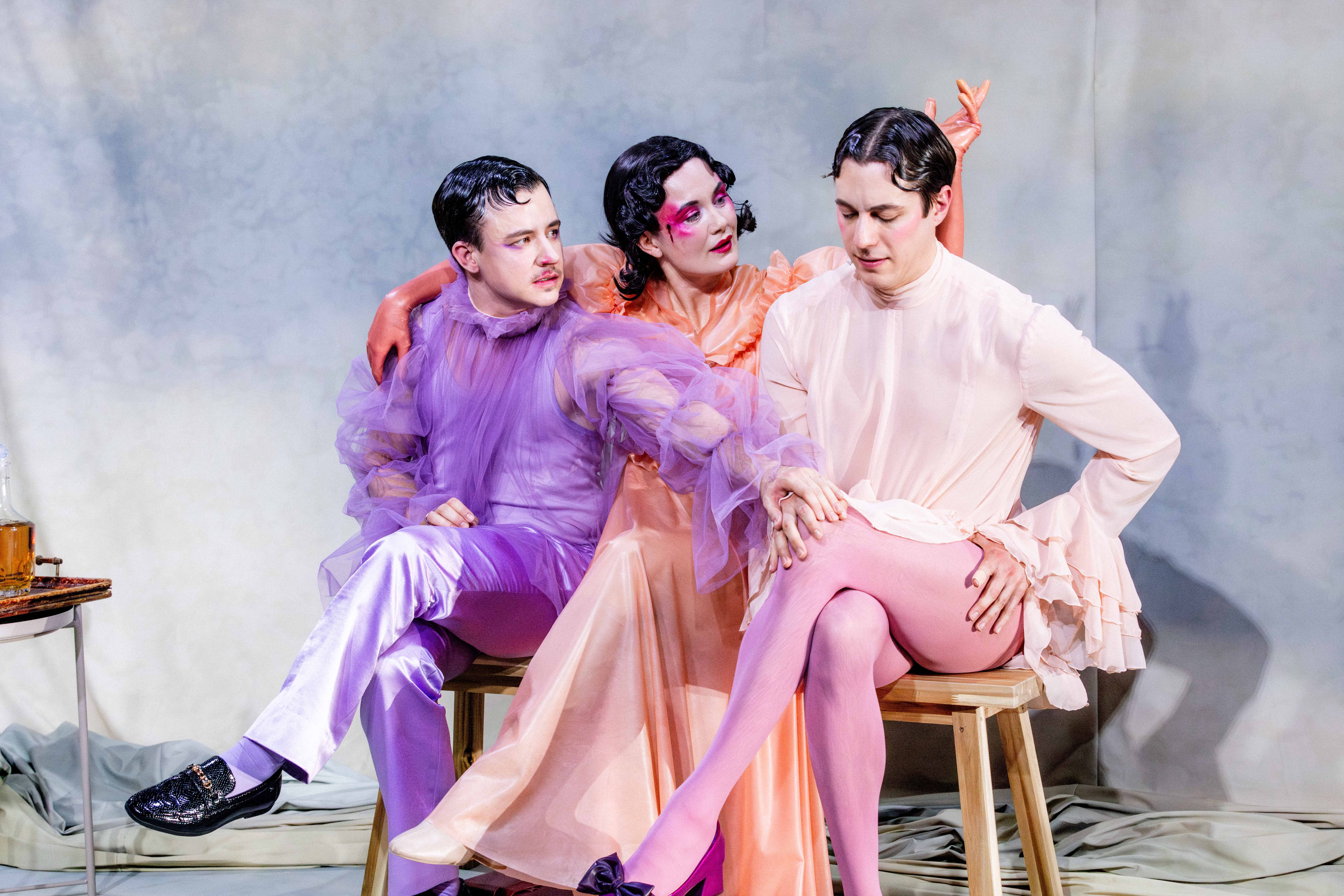 Tre personer i fargerike scenekostymer sitter tett sammen på en benk i et teatralt oppstilt rom