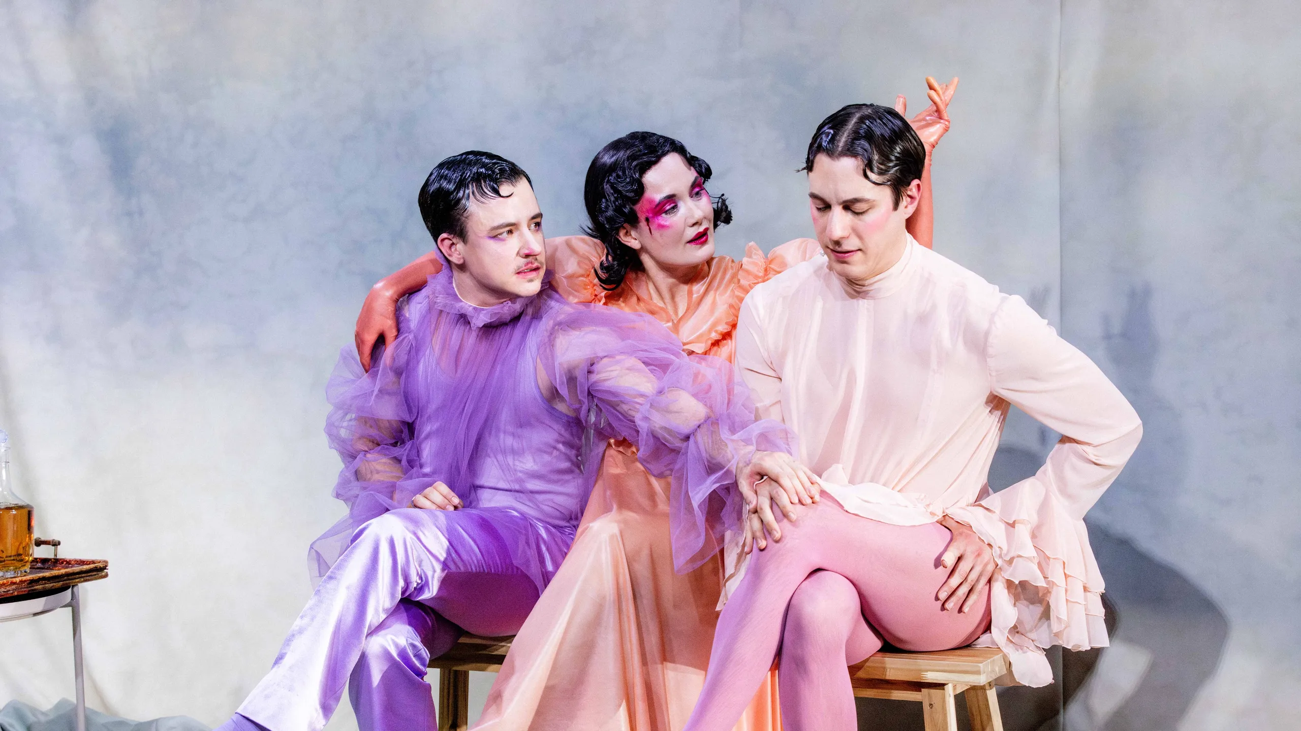 Tre personer i fargerike scenekostymer sitter tett sammen på en benk i et teatralt oppstilt rom