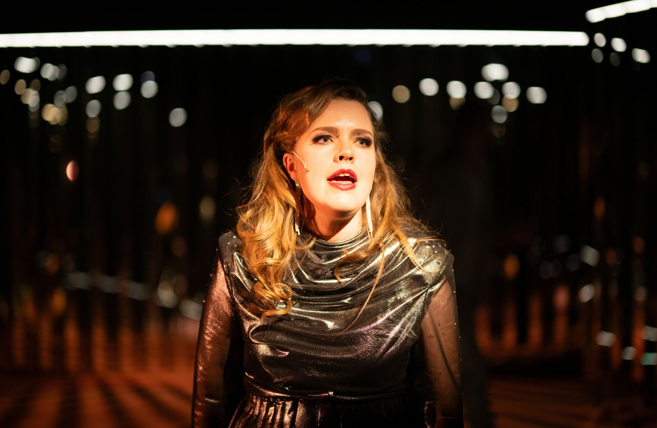 Kvinne i glitrende mørk kjole og langt hår står i scenelys og synger, med mørkt rom og lys i bakgrunnen.