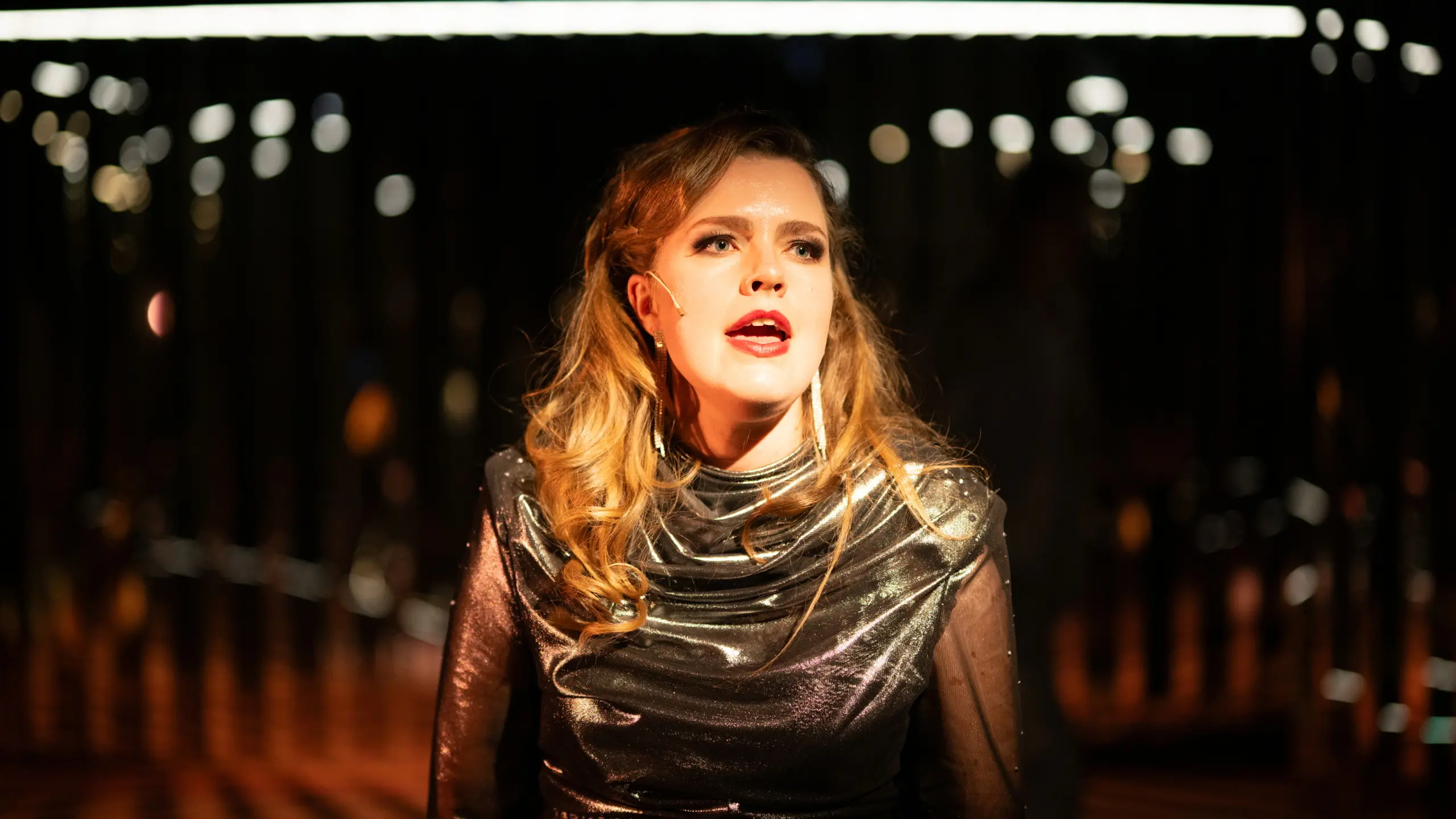 Kvinne i glitrende mørk kjole og langt hår står i scenelys og synger, med mørkt rom og lys i bakgrunnen.