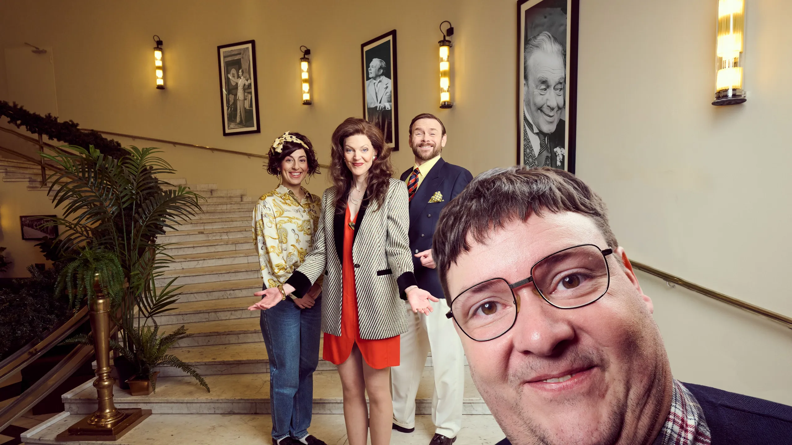 tre personer i fargerike kostymer poserer smilende i en trapp med marmor og palmer, mens én mann står tett foran kamera med briller og rød sløyfe.