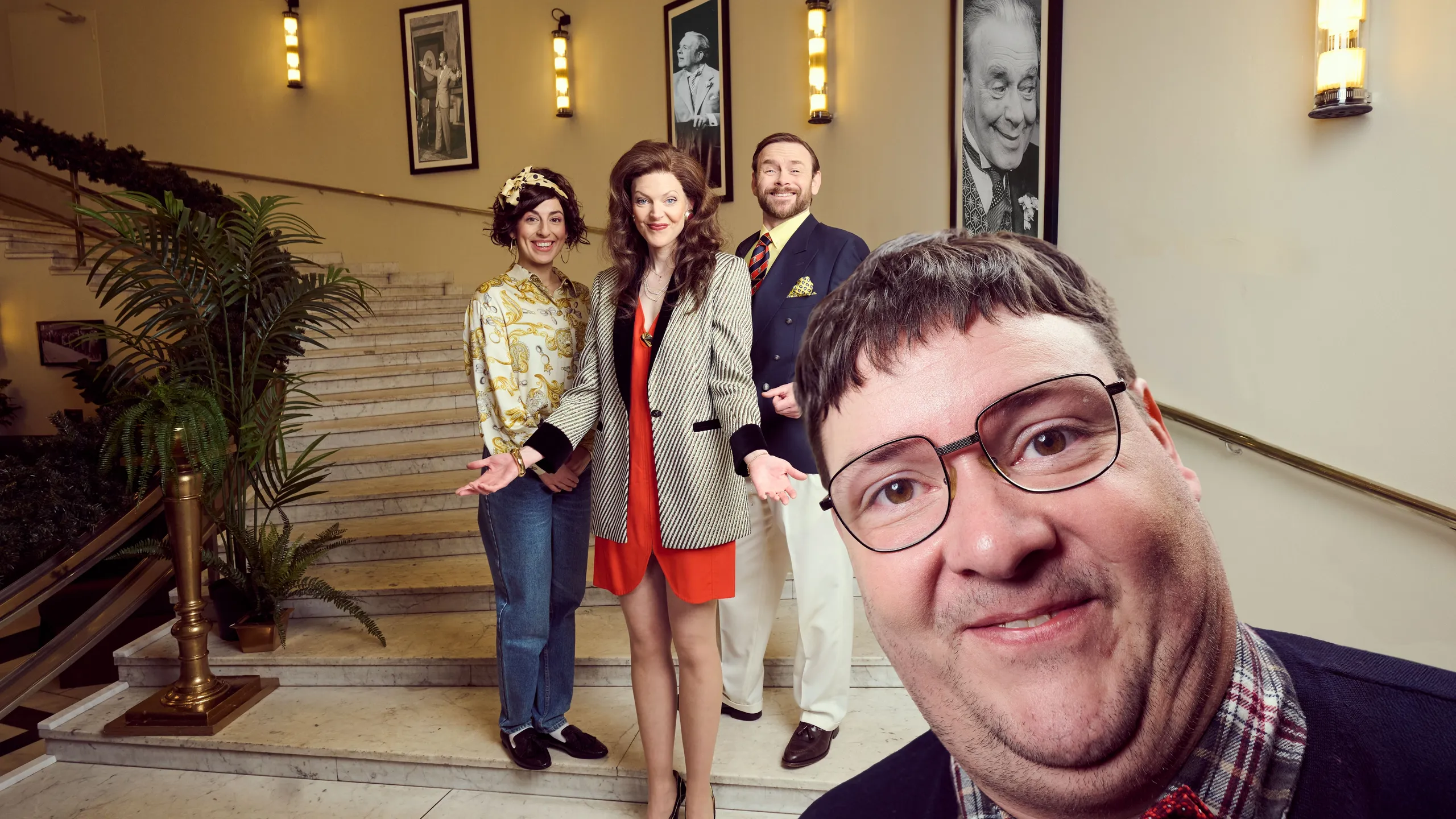 tre personer i fargerike kostymer poserer smilende i en trapp med marmor og palmer, mens én mann står tett foran kamera med briller og rød sløyfe.