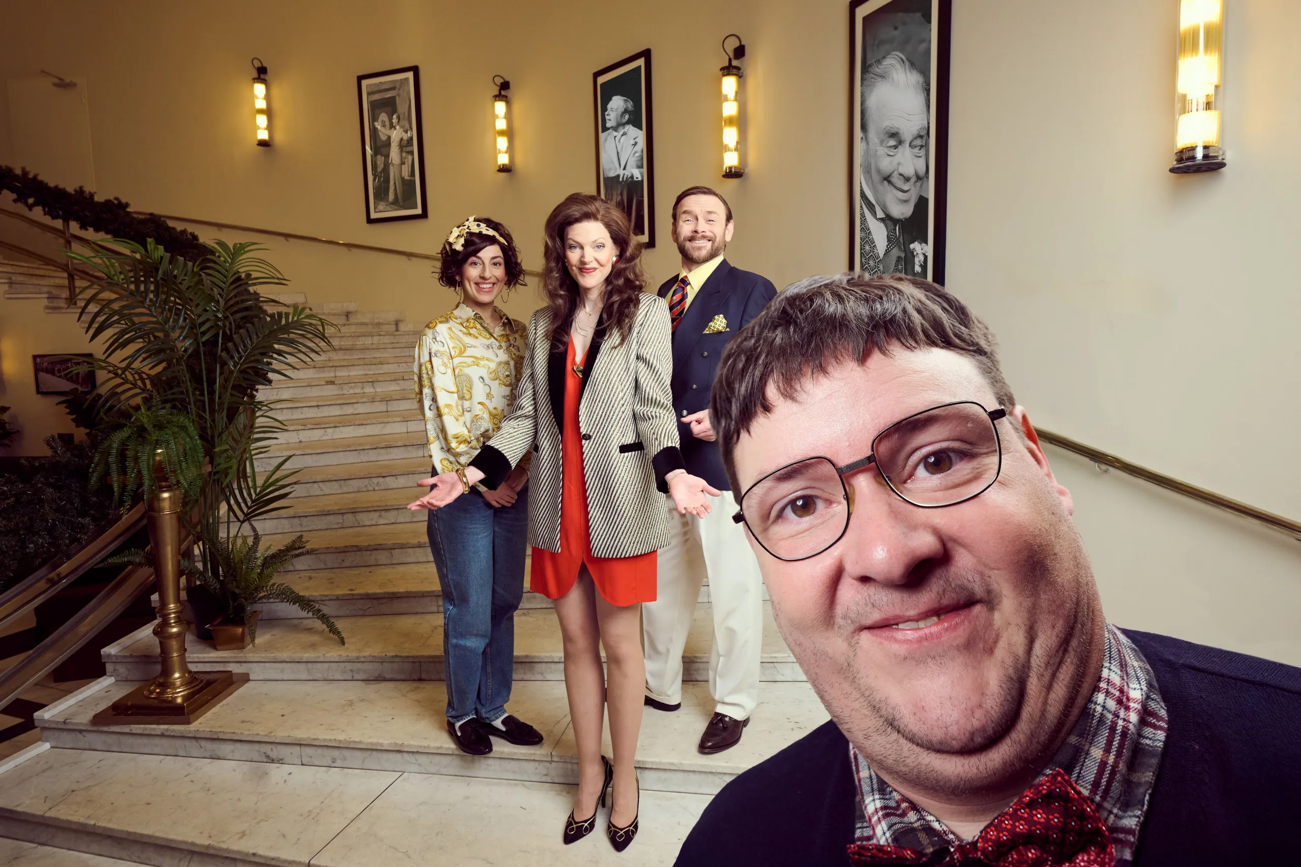 tre personer i fargerike kostymer poserer smilende i en trapp med marmor og palmer, mens én mann står tett foran kamera med briller og rød sløyfe.