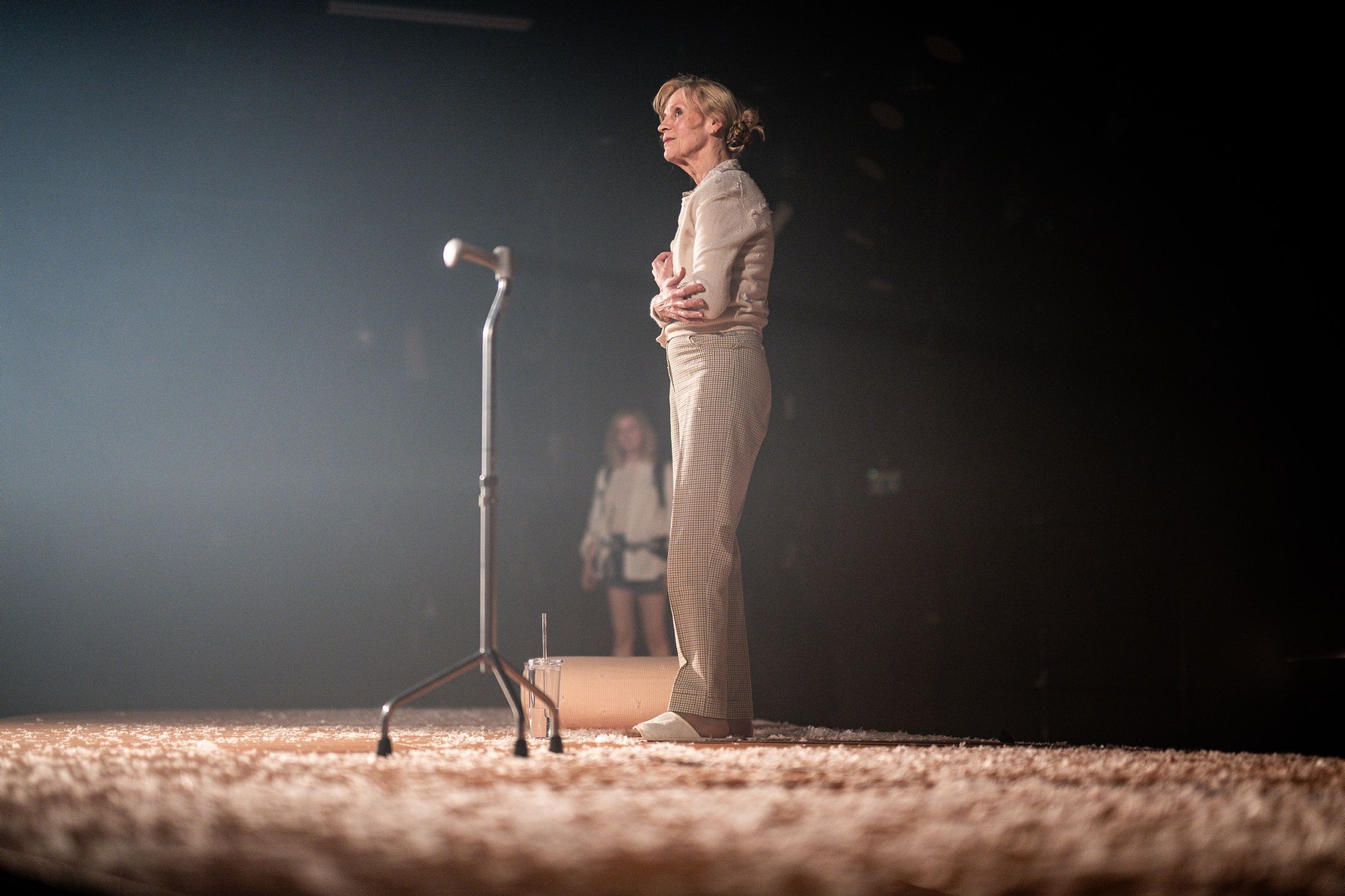 kvinne står alene på teaterscene ved et stativ, omgitt av mykt scenelys og hvitt gulvdekke