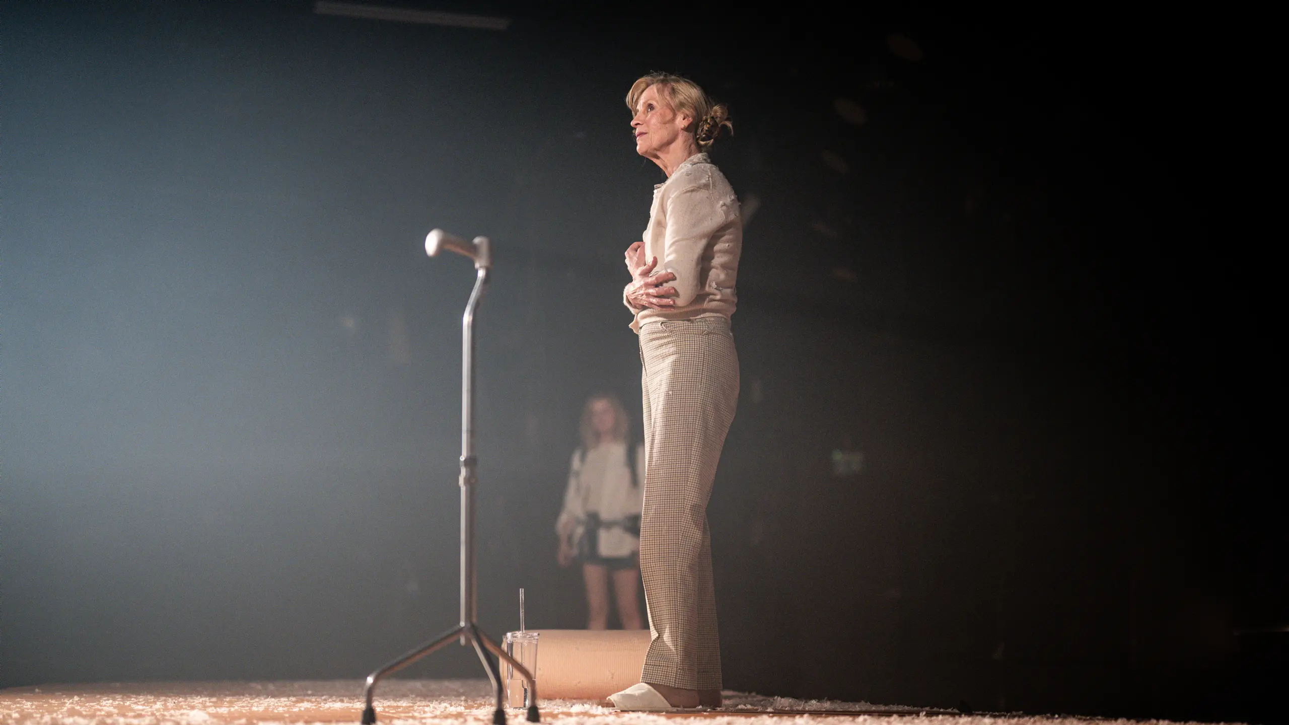 kvinne står alene på teaterscene ved et stativ, omgitt av mykt scenelys og hvitt gulvdekke
