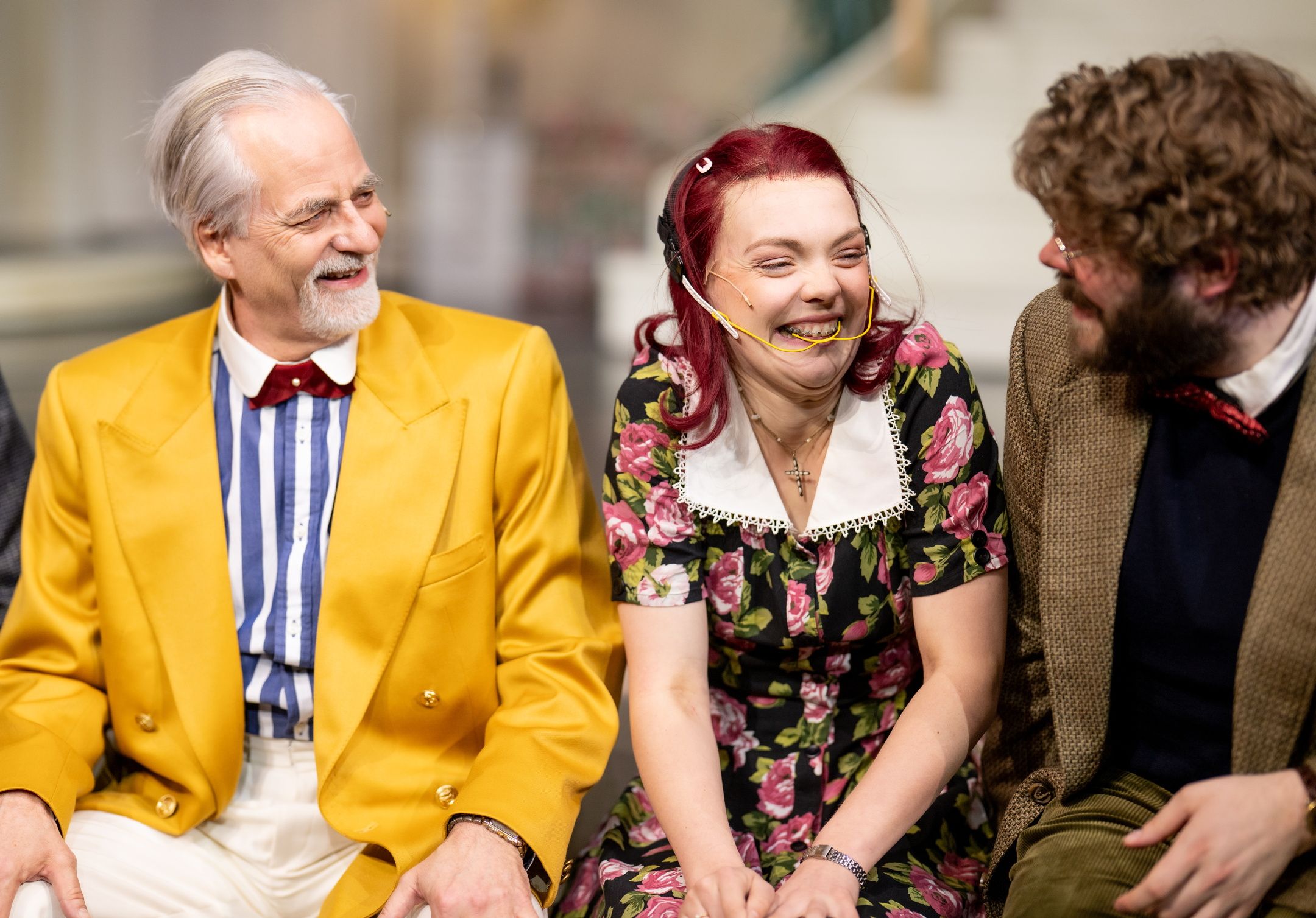 Tre personer sitter tett sammen og ler på teaterscene, én i gul blazer og én i blomsterkjole