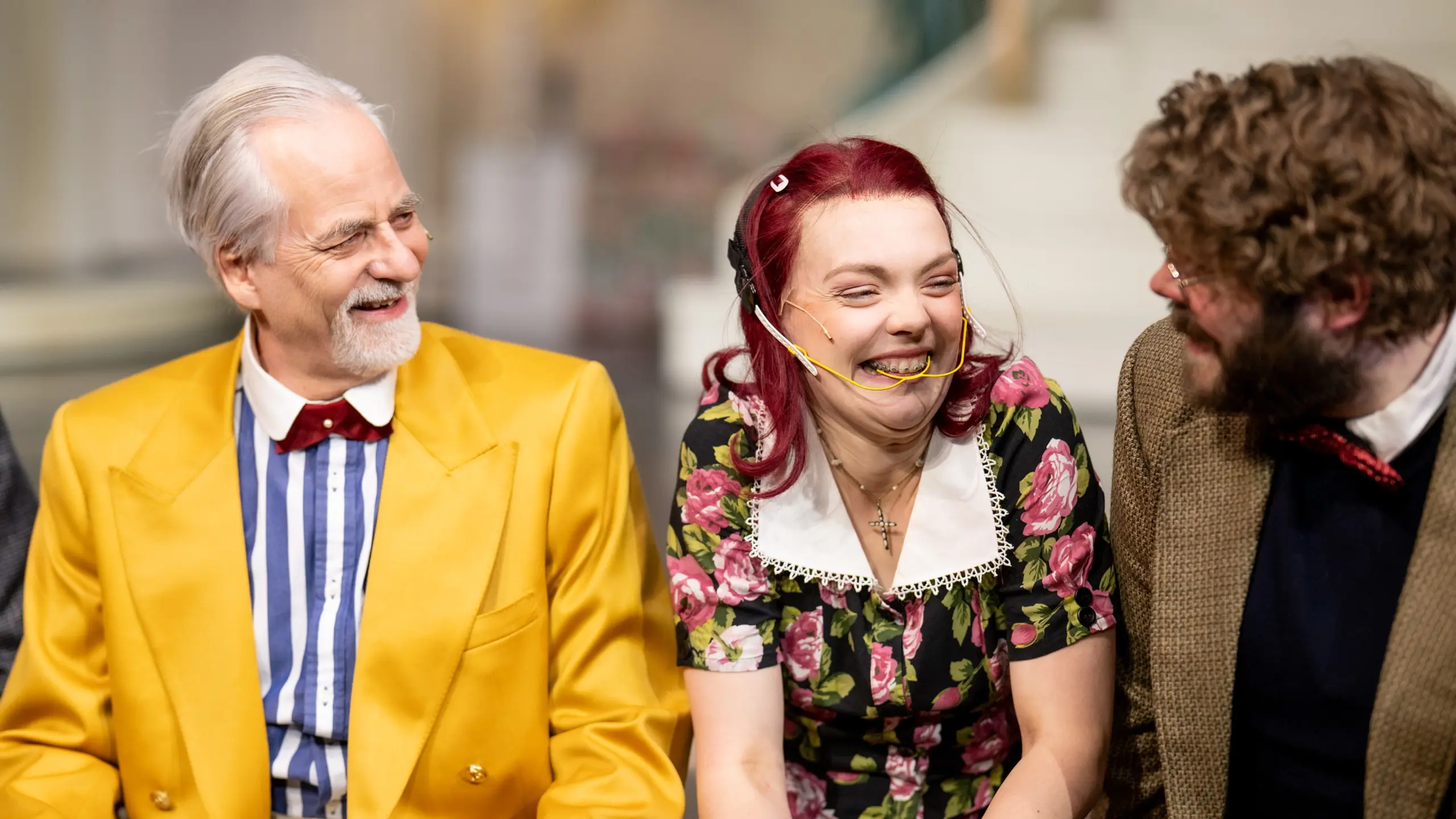 Tre personer sitter tett sammen og ler på teaterscene, én i gul blazer og én i blomsterkjole