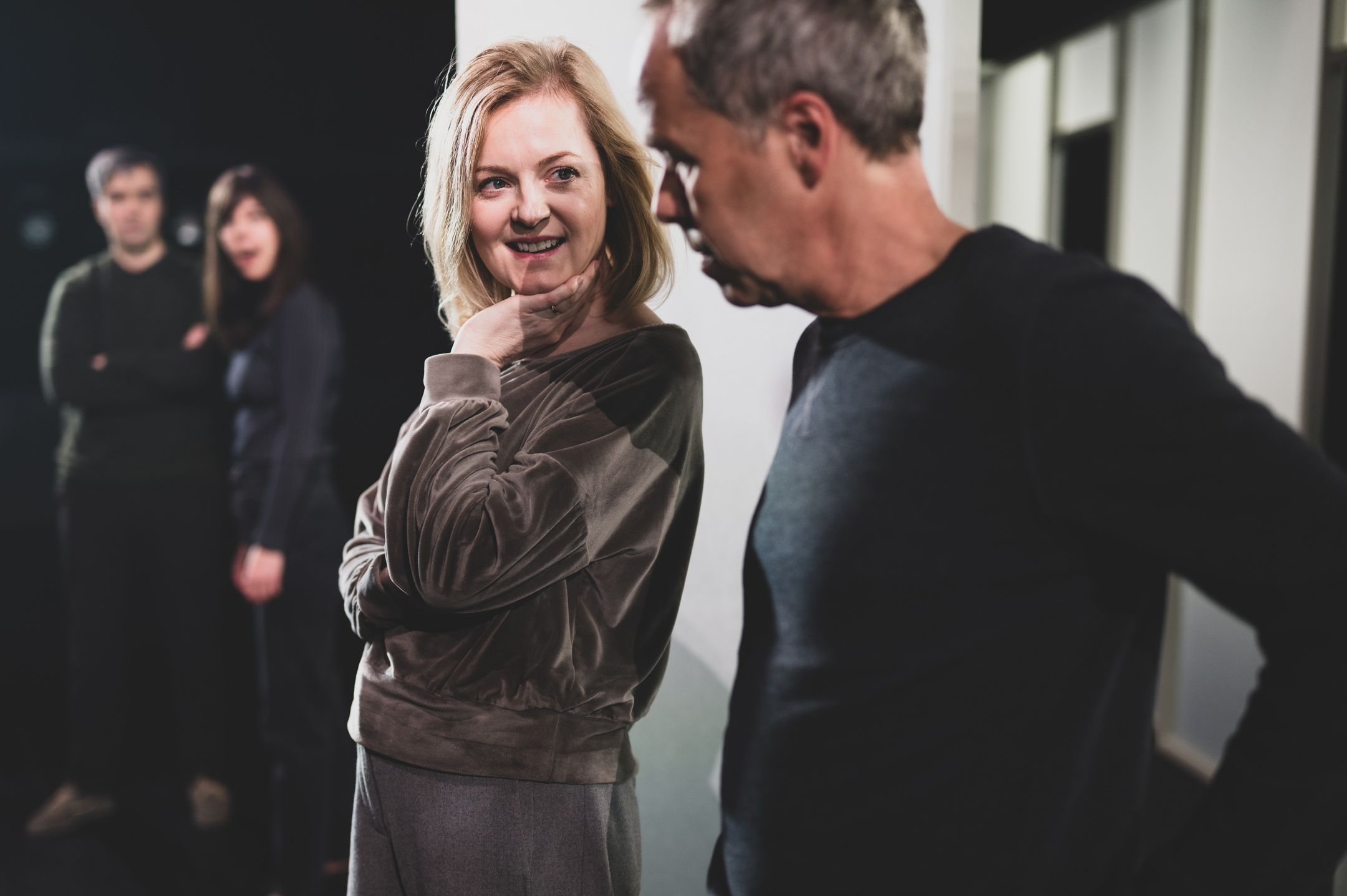 En kvinne smiler og holder hånden lett under haken mens hun ser på en mann som står ved siden av henne, med to personer uskarpt i bakgrunnen.
