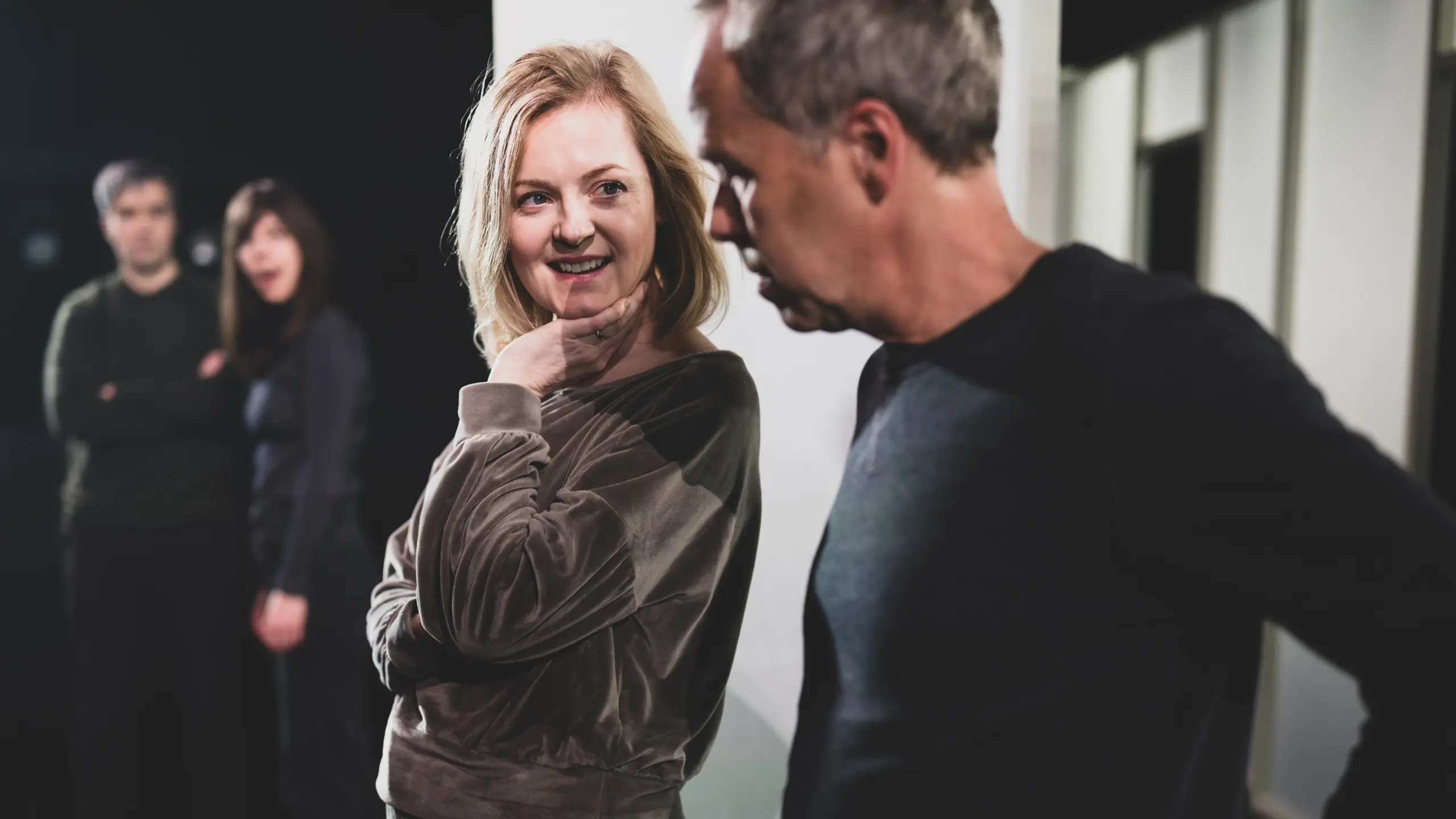 En kvinne smiler og holder hånden lett under haken mens hun ser på en mann som står ved siden av henne, med to personer uskarpt i bakgrunnen.