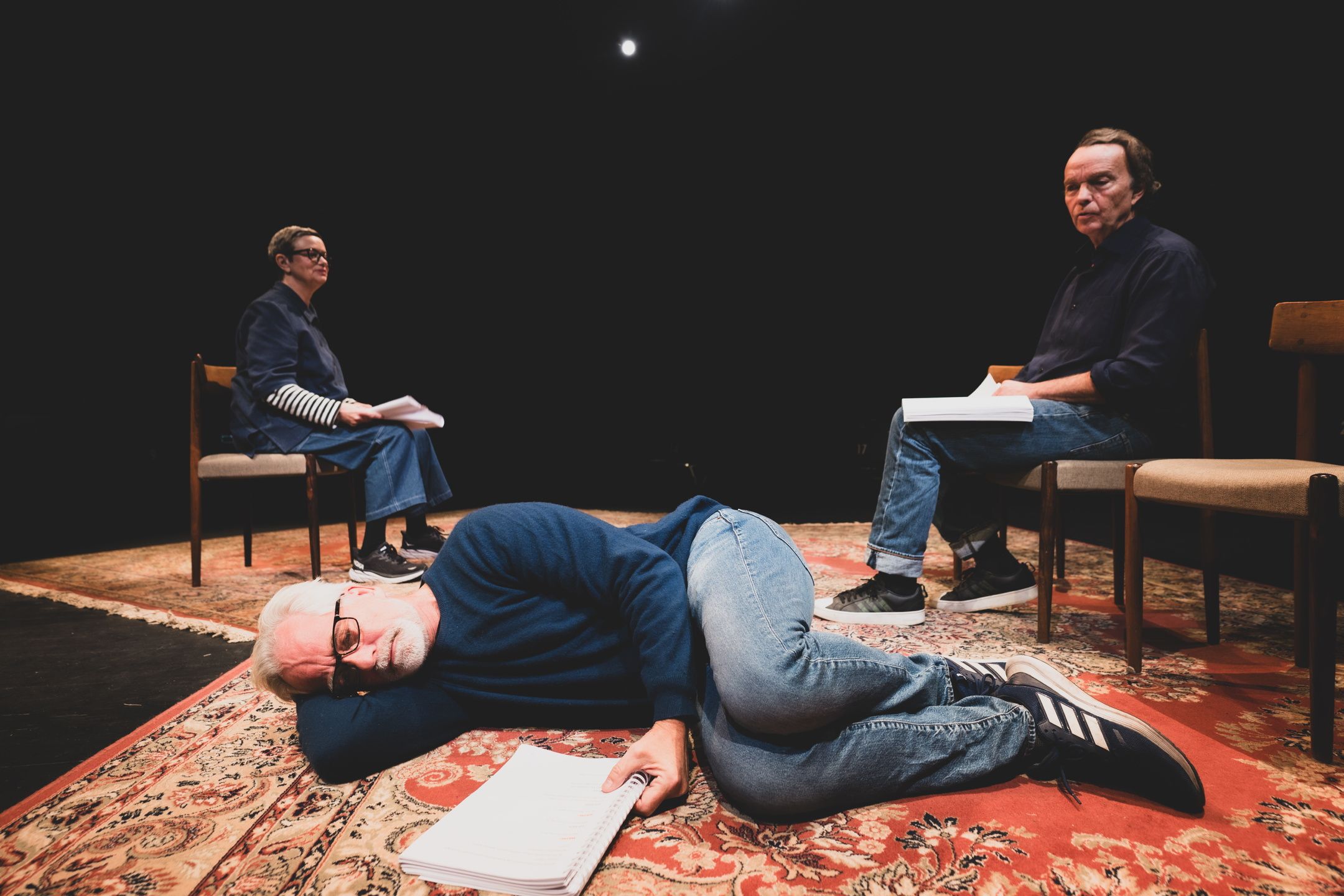 En mann ligger på siden på et teppe med et manus foran seg. Bak sitter en mann og en kvinne på stoler med manus på fanger