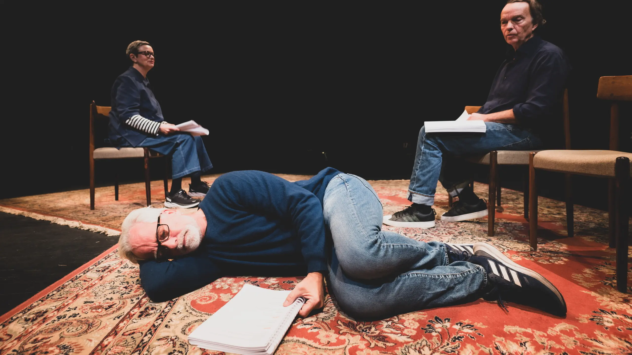 En mann ligger på siden på et teppe med et manus foran seg. Bak sitter en mann og en kvinne på stoler med manus på fanger