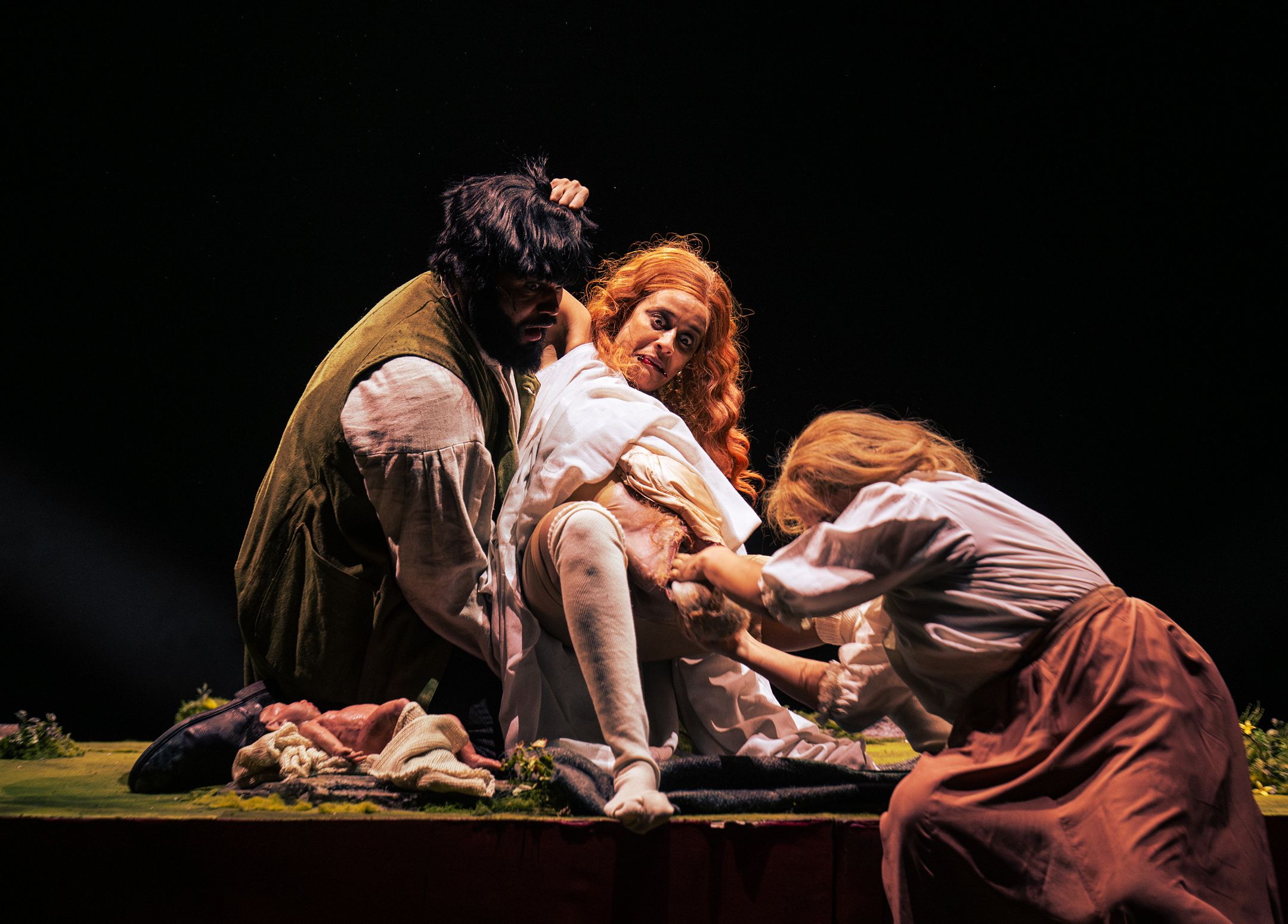 Tre skuespillere i historiske kostymer spiller ut en intens fødselsscene med dramatisk belysning og kroppslig innsats.
