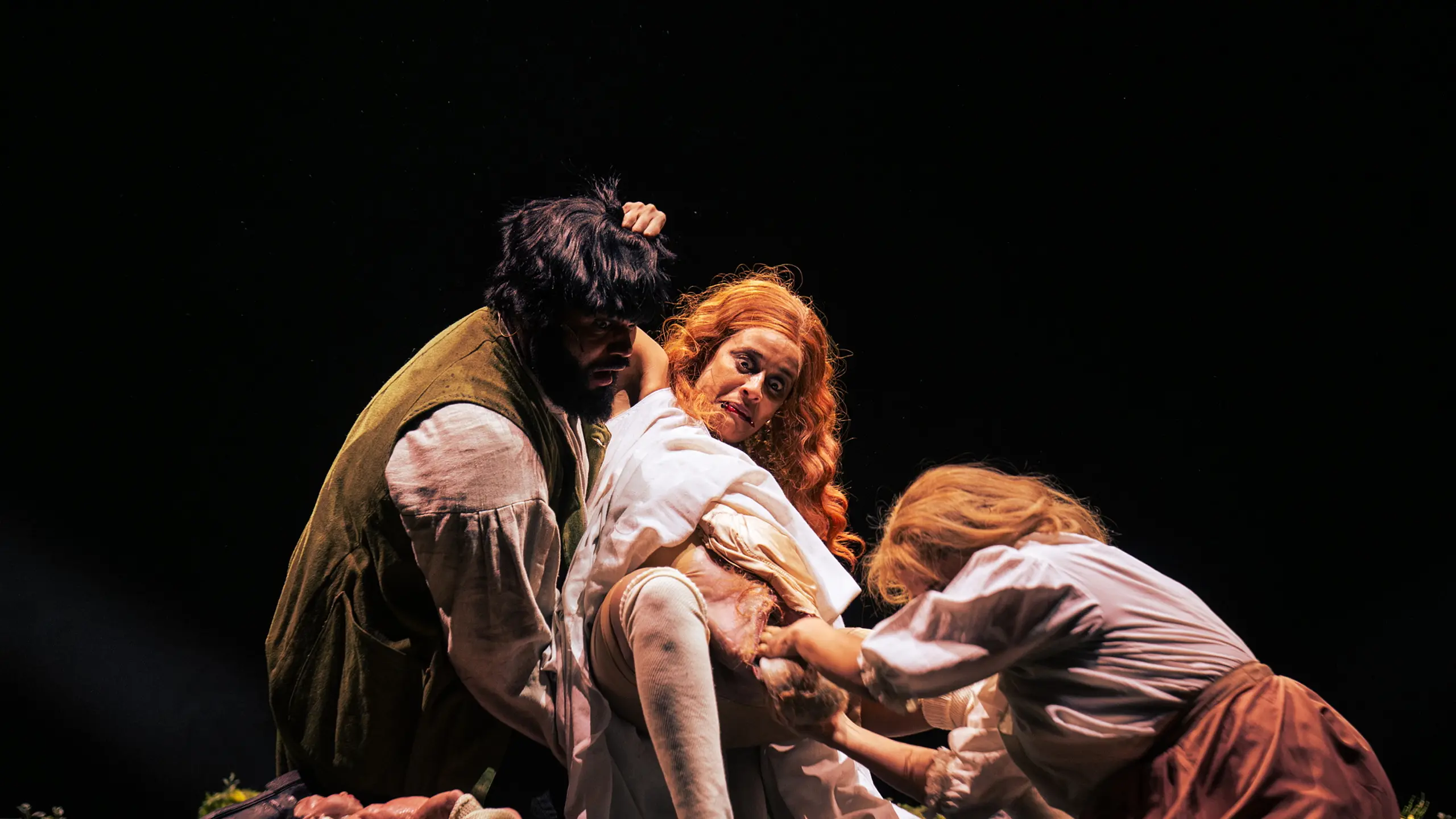 Tre skuespillere i historiske kostymer spiller ut en intens fødselsscene med dramatisk belysning og kroppslig innsats.