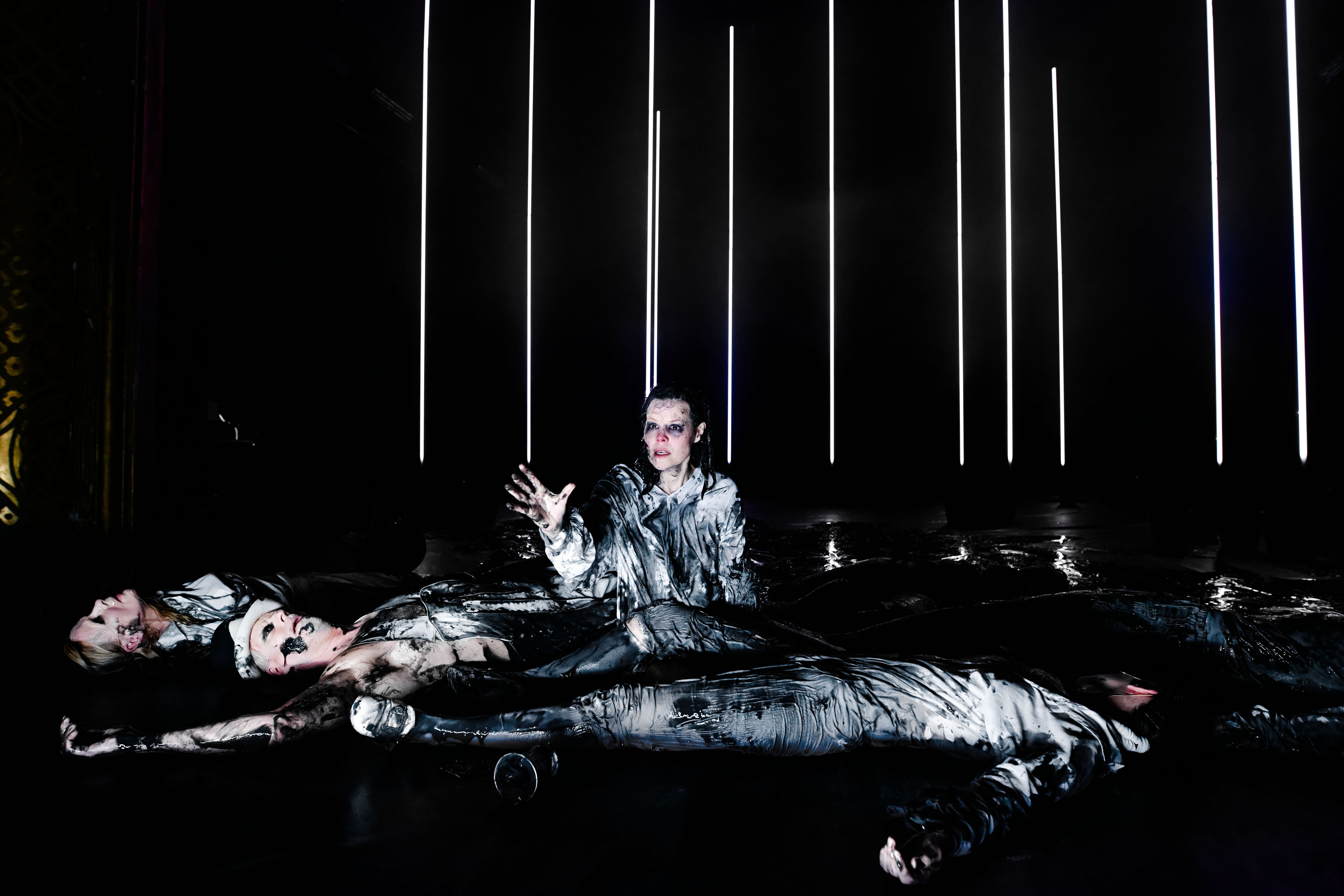 En kvinne sitter på mørk scene med lyskjegler. Rundt henne ligger flere skuespillere. Alle er tilgriset i svart klissete væske