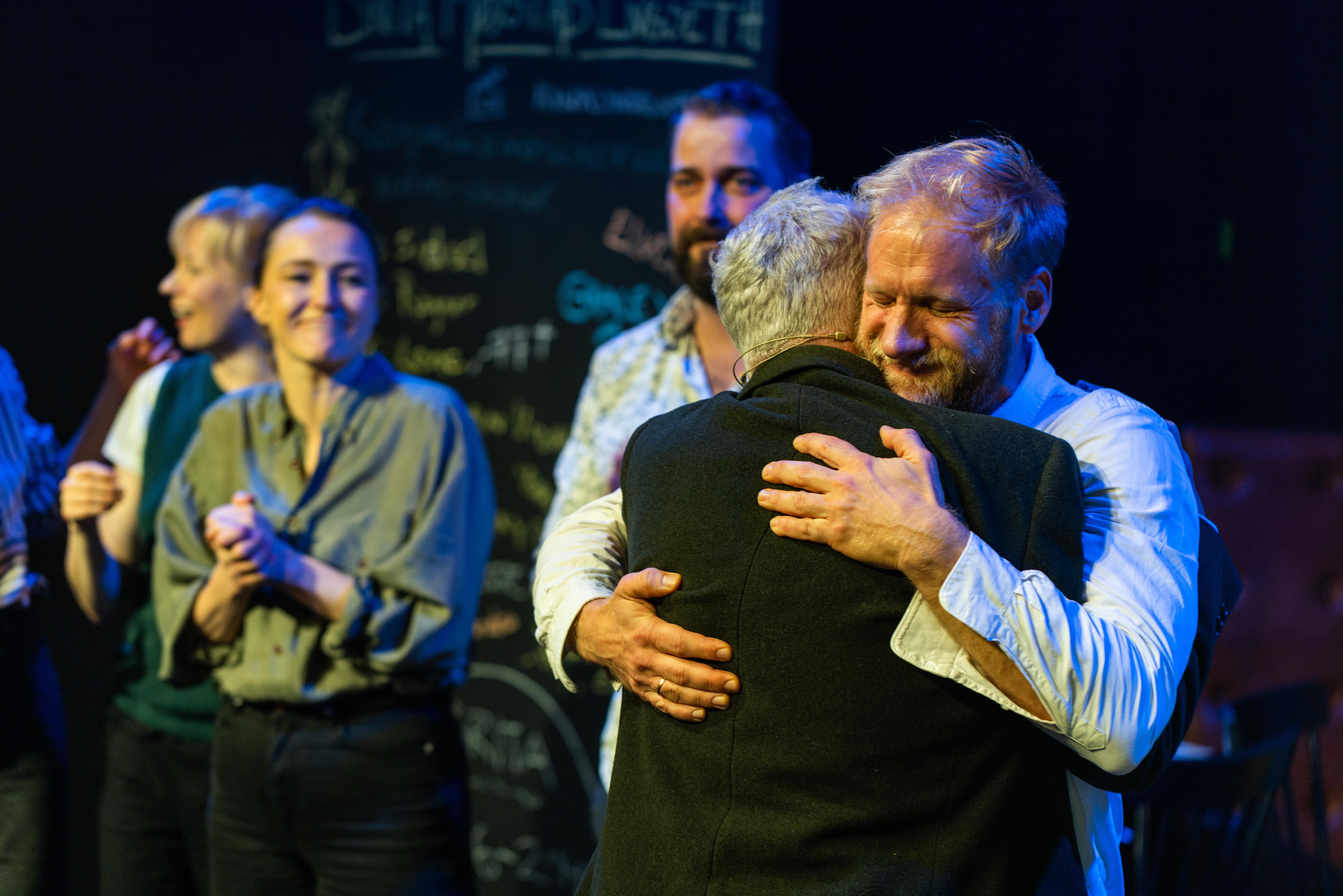 To personer omfavner hverandre varmt på scene, andre smiler og klapper i bakgrunnen