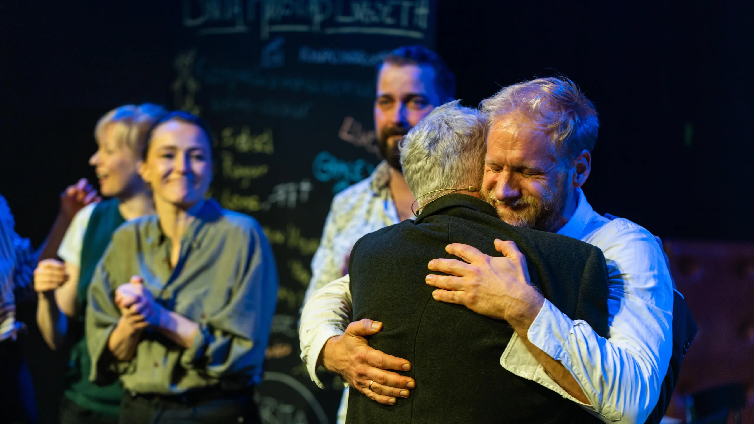 To personer omfavner hverandre varmt på scene, andre smiler og klapper i bakgrunnen