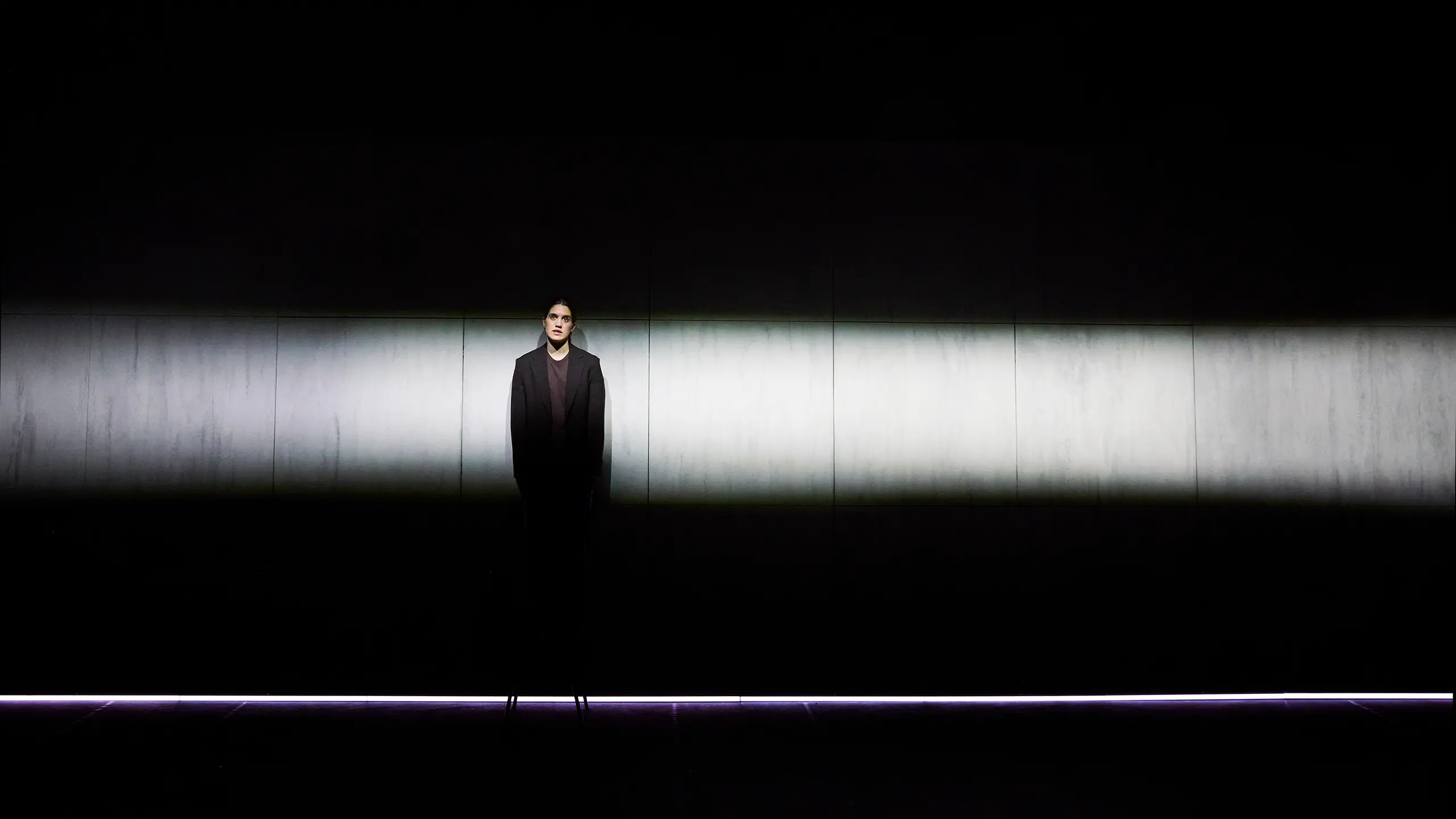 En kvinne i sort blazer står under en smal lysstripe på en mørklagt scene, omringet av mørke.