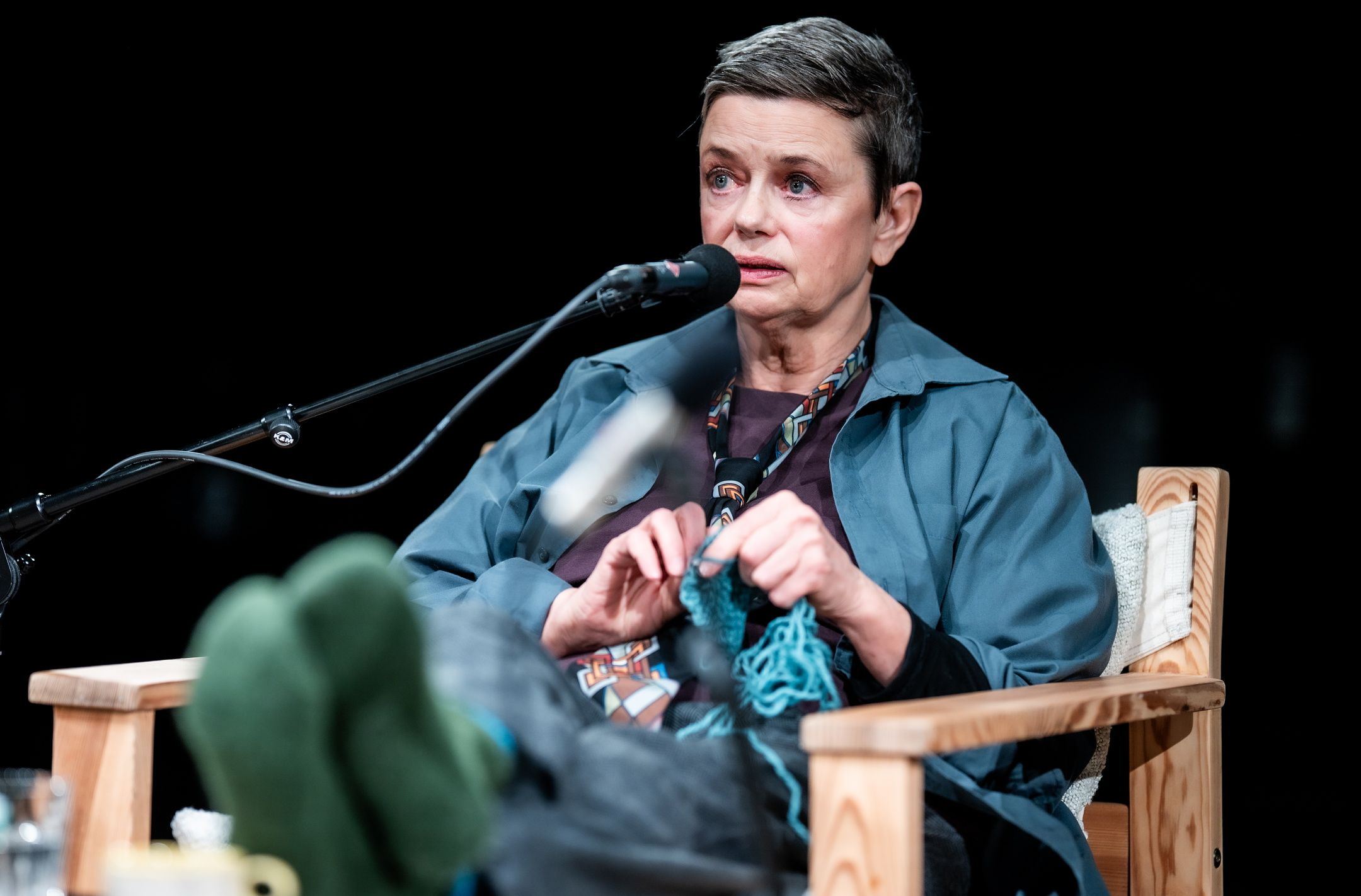 Person med kort hår sitter i stol med mikrofon og strikker blått garn mens hun snakker.