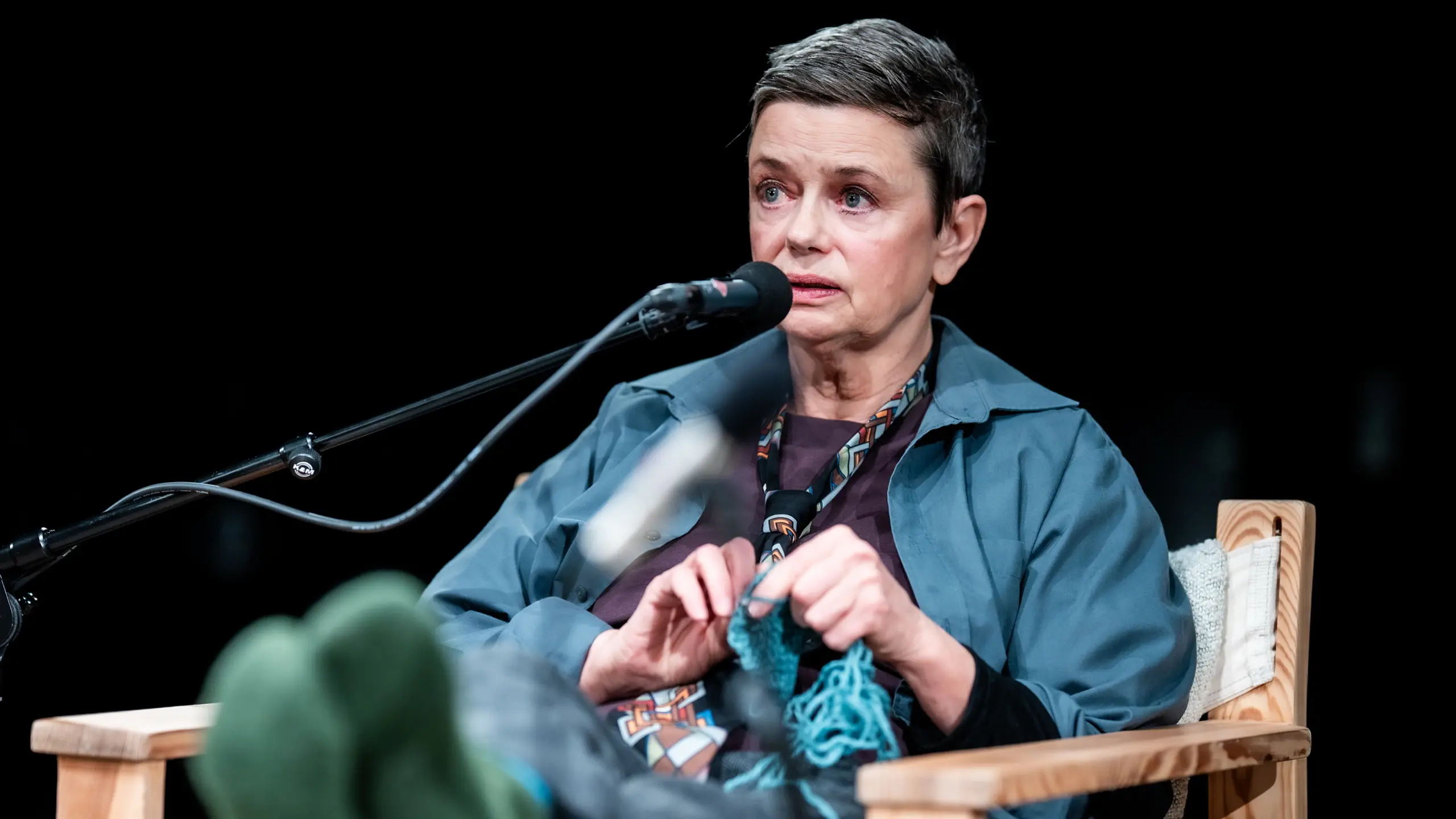 Person med kort hår sitter i stol med mikrofon og strikker blått garn mens hun snakker.