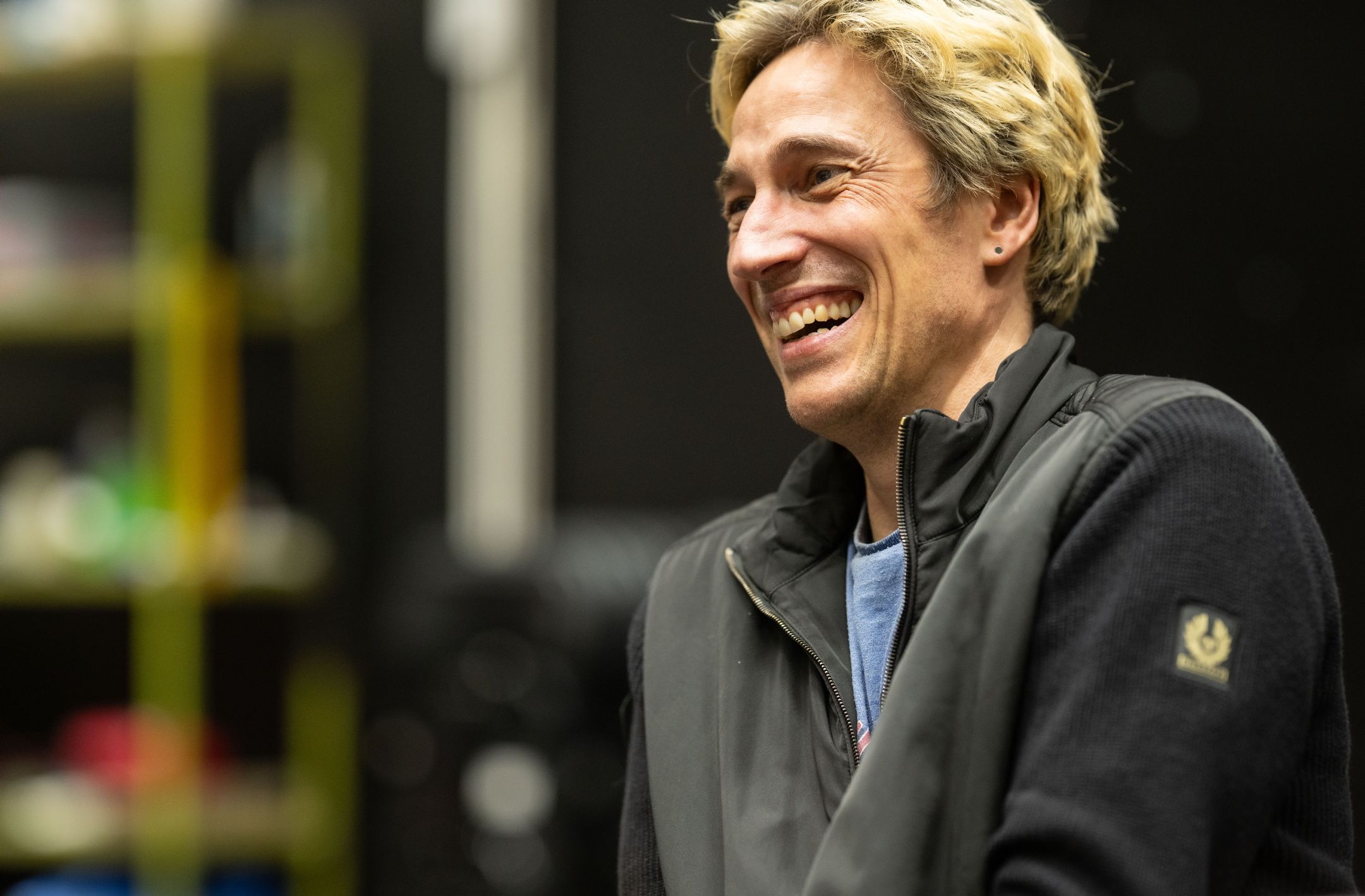 En mann med lyst hår og sort jakke ler under en prøve med rekvisitter og sort bakgrunn.