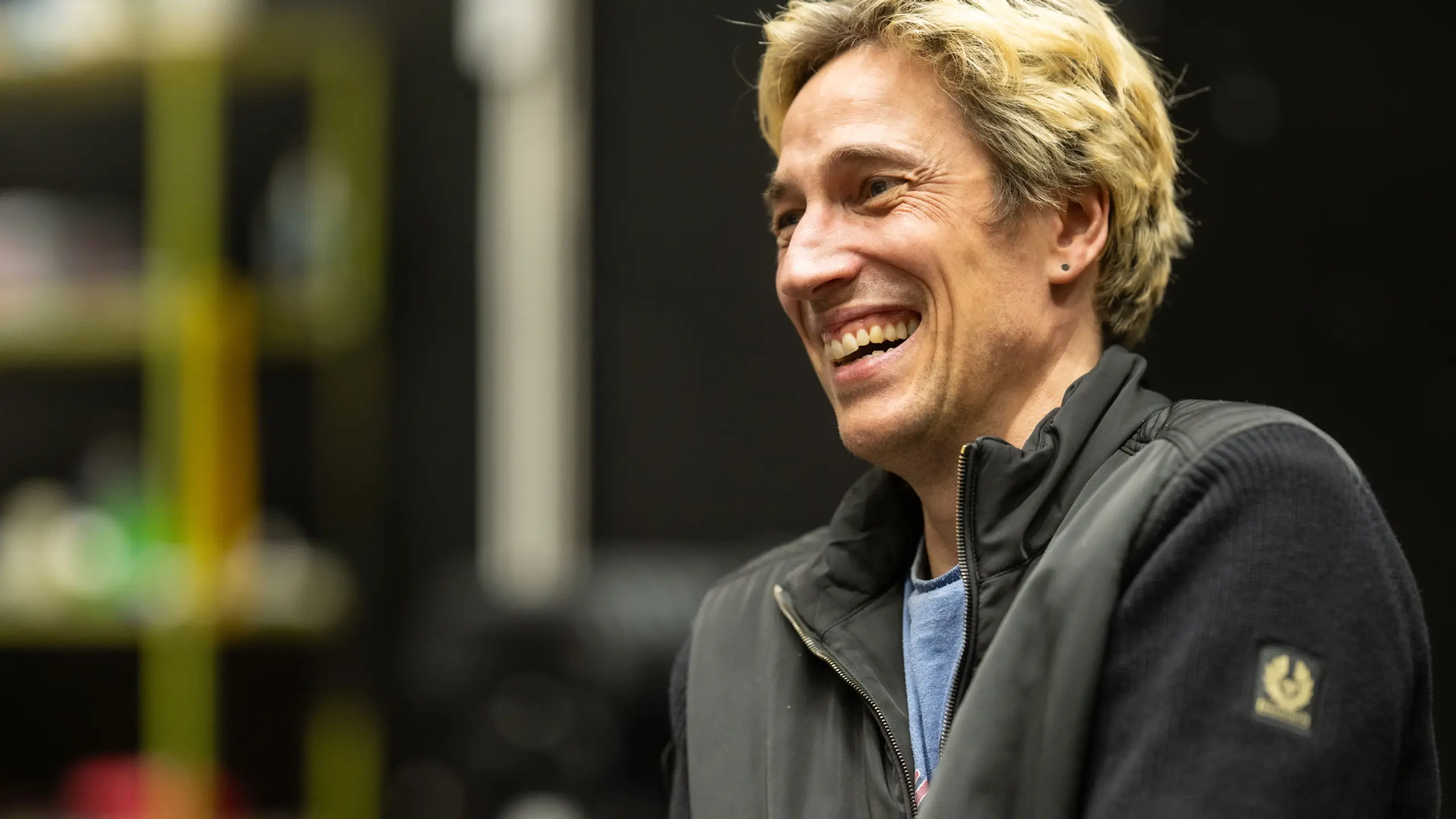 En mann med lyst hår og sort jakke ler under en prøve med rekvisitter og sort bakgrunn.