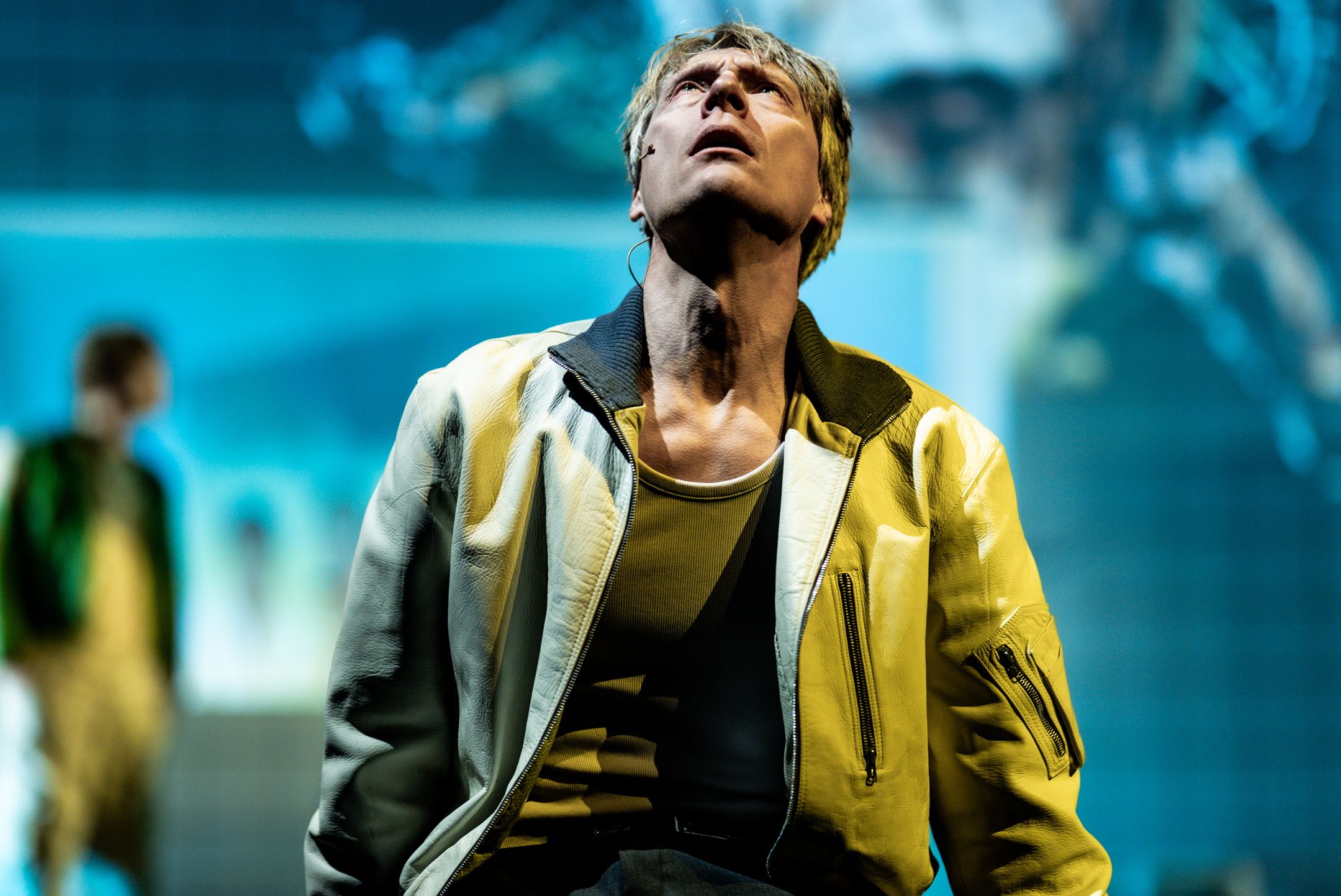 Mann i gul jakke ser opp med alvorlig uttrykk, mot uskarp blå bakgrunn og med mikrofon på kinnet.