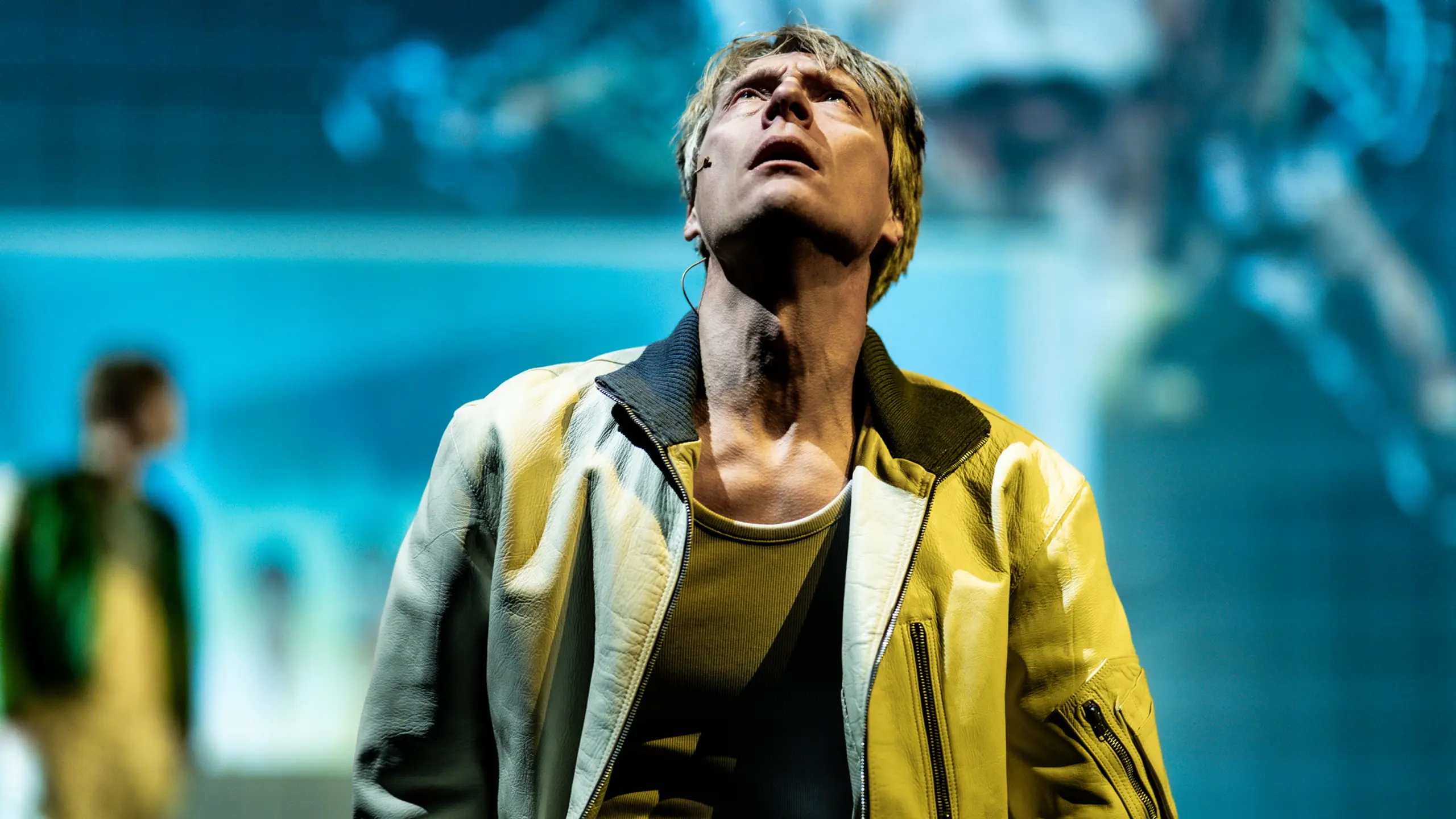 Mann i gul jakke ser opp med alvorlig uttrykk, mot uskarp blå bakgrunn og med mikrofon på kinnet.