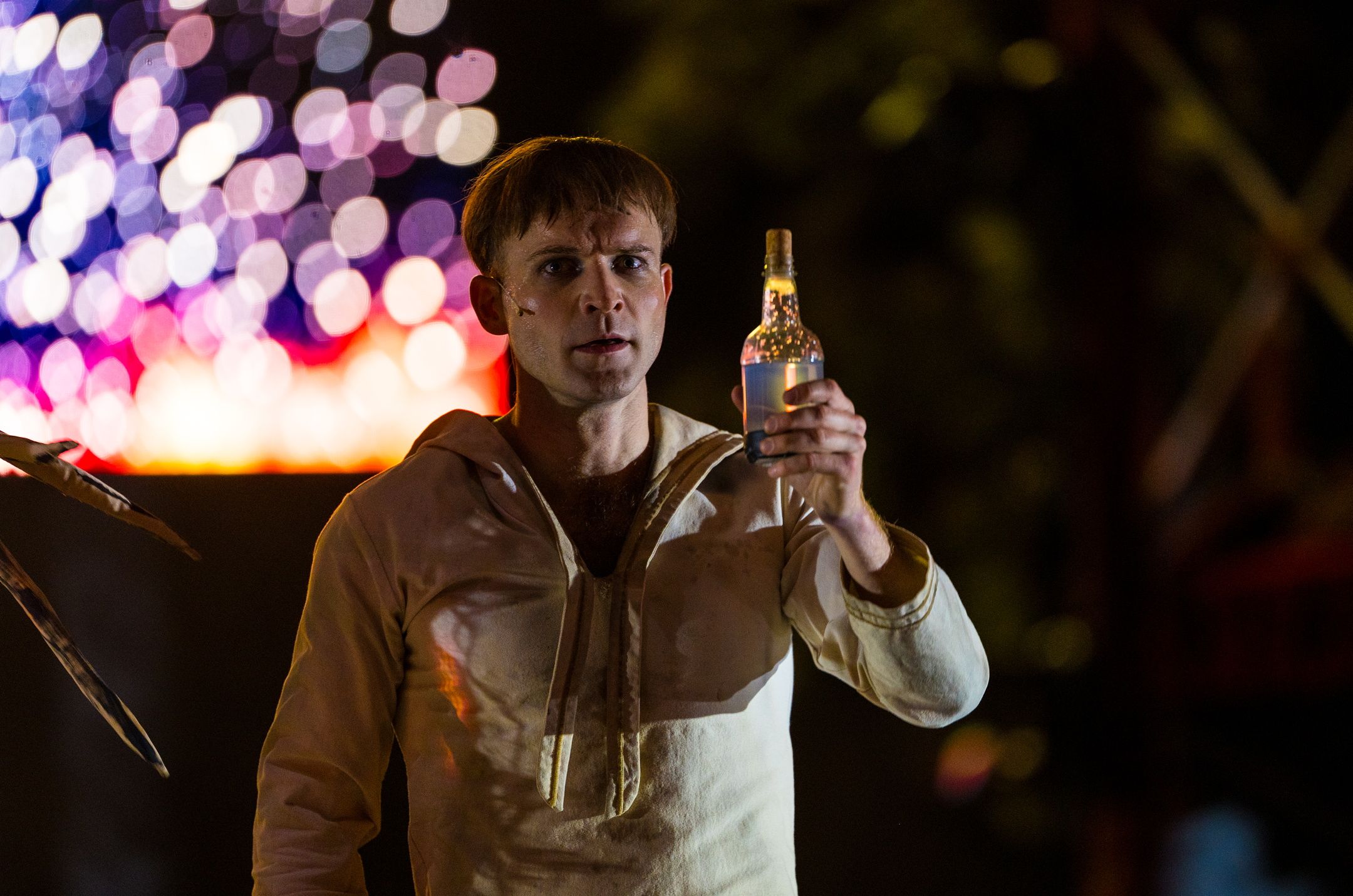 Skuespiller i hvit drakt holder opp en glassflaske i scenelys med fargerike lysrefleksjoner i bakgrunnen.