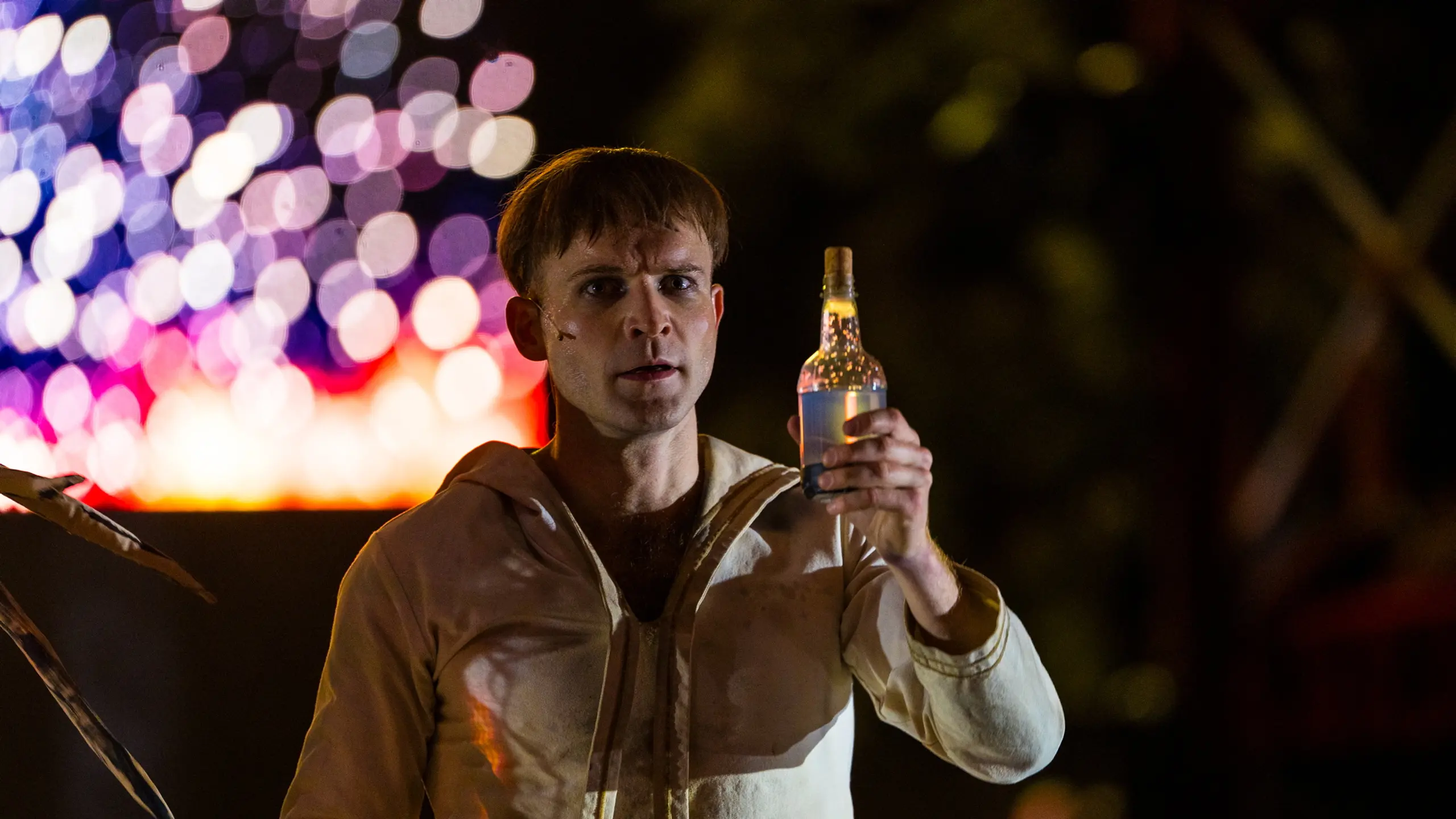 Skuespiller i hvit drakt holder opp en glassflaske i scenelys med fargerike lysrefleksjoner i bakgrunnen.